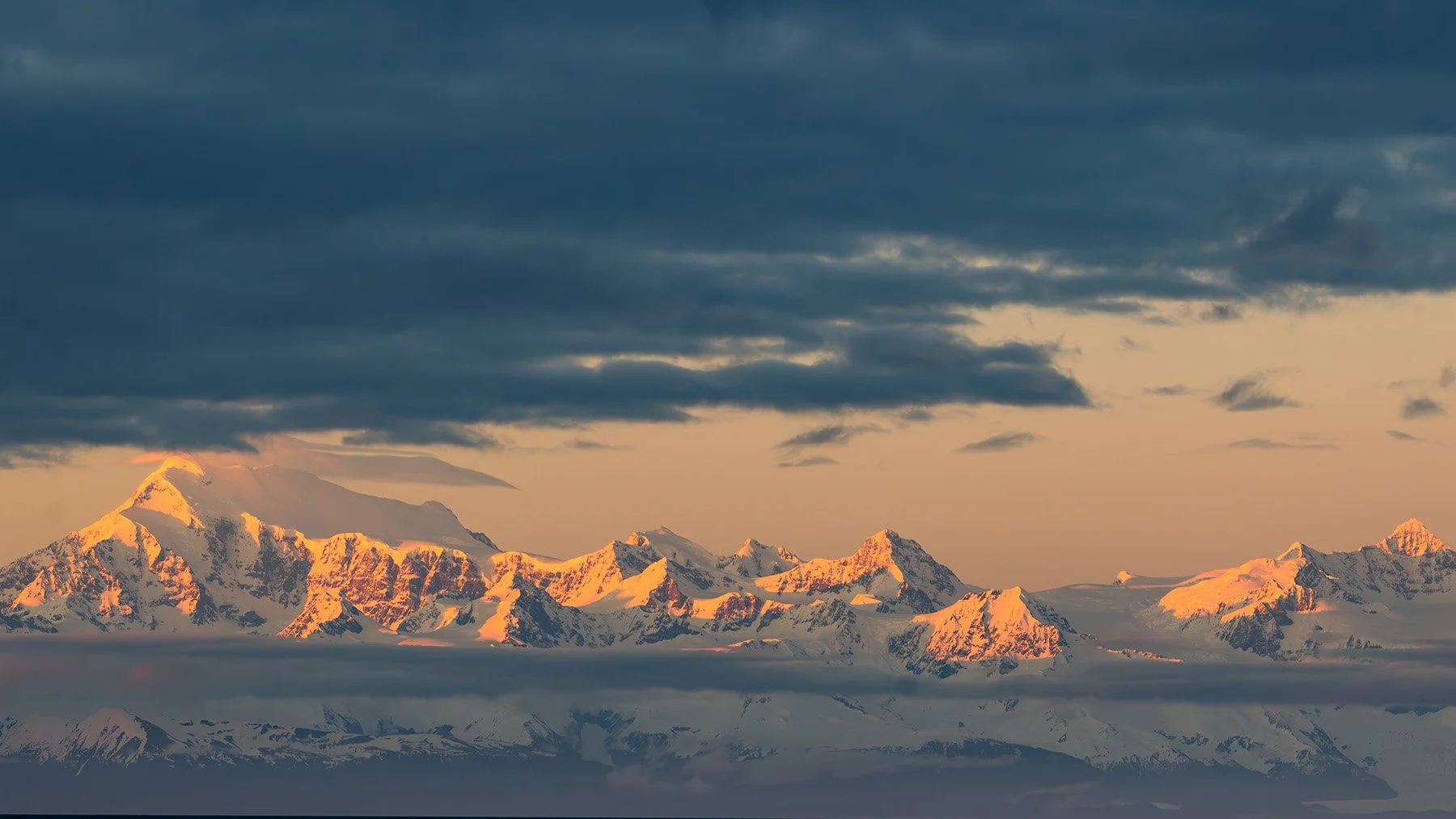 Snow-covered mountain range at sunset with dramatic clouds.