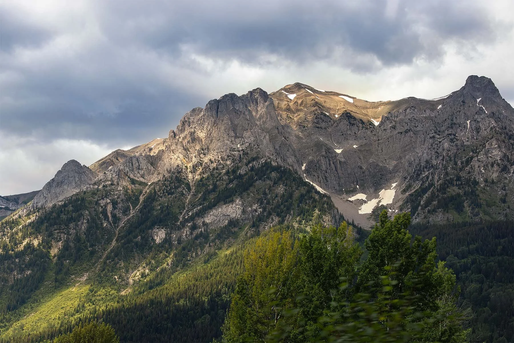 Panoramic view of a rugged mountain range with patches of snow and lush green forests under a cloudy sky.