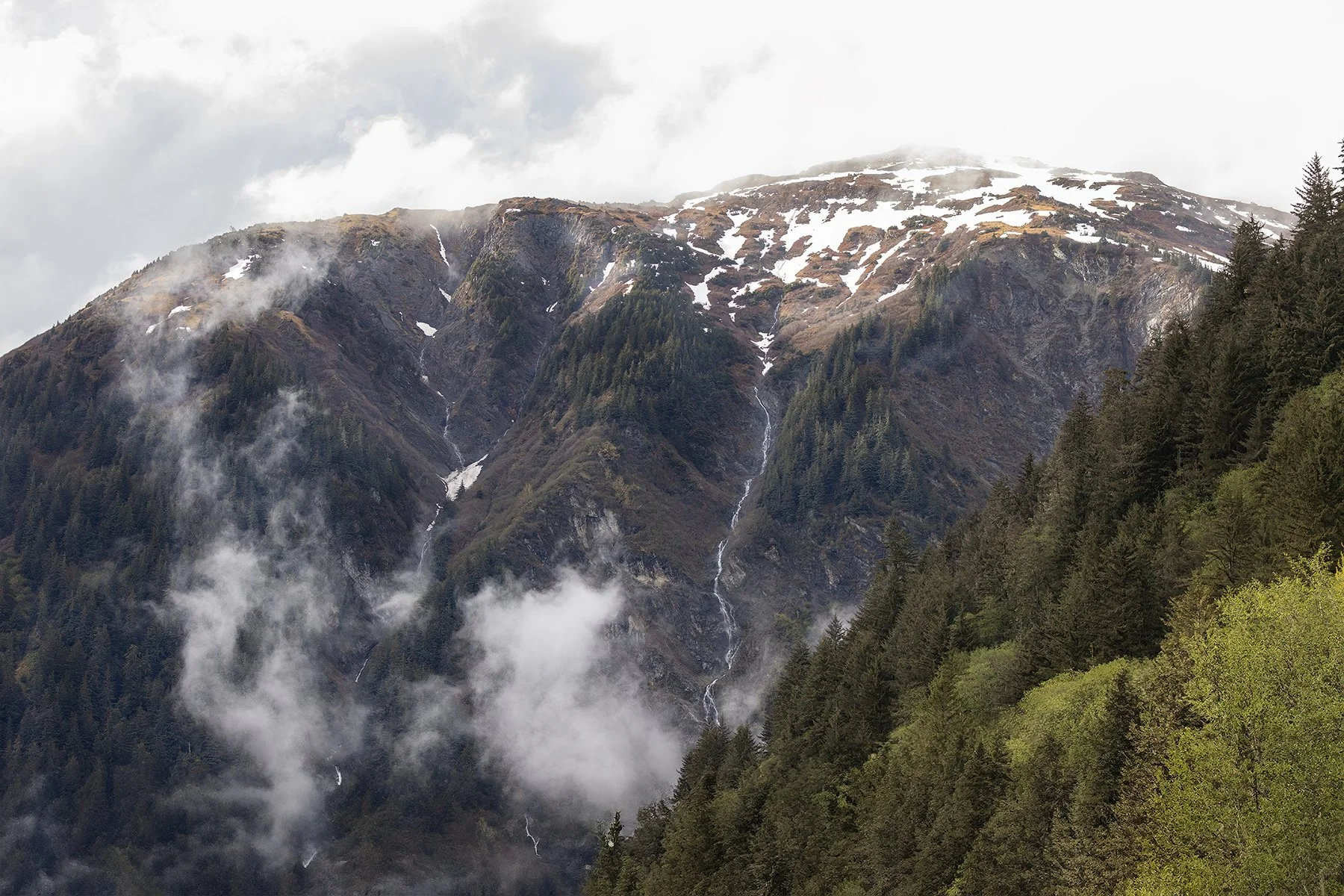 Mountain landscape with snow patches, forested slopes, and low clouds.