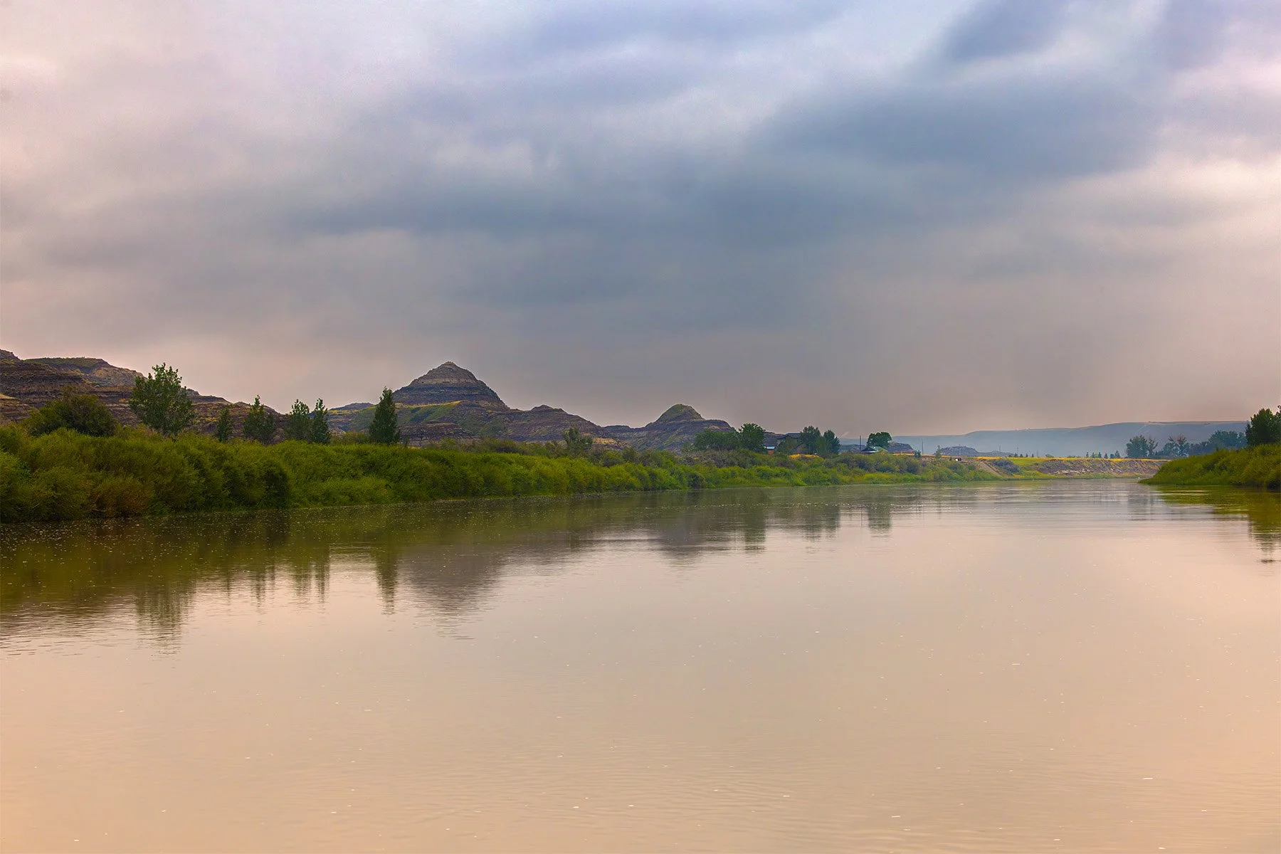 River with calm water, surrounded by green shrubbery and distant hills under a cloudy sky.