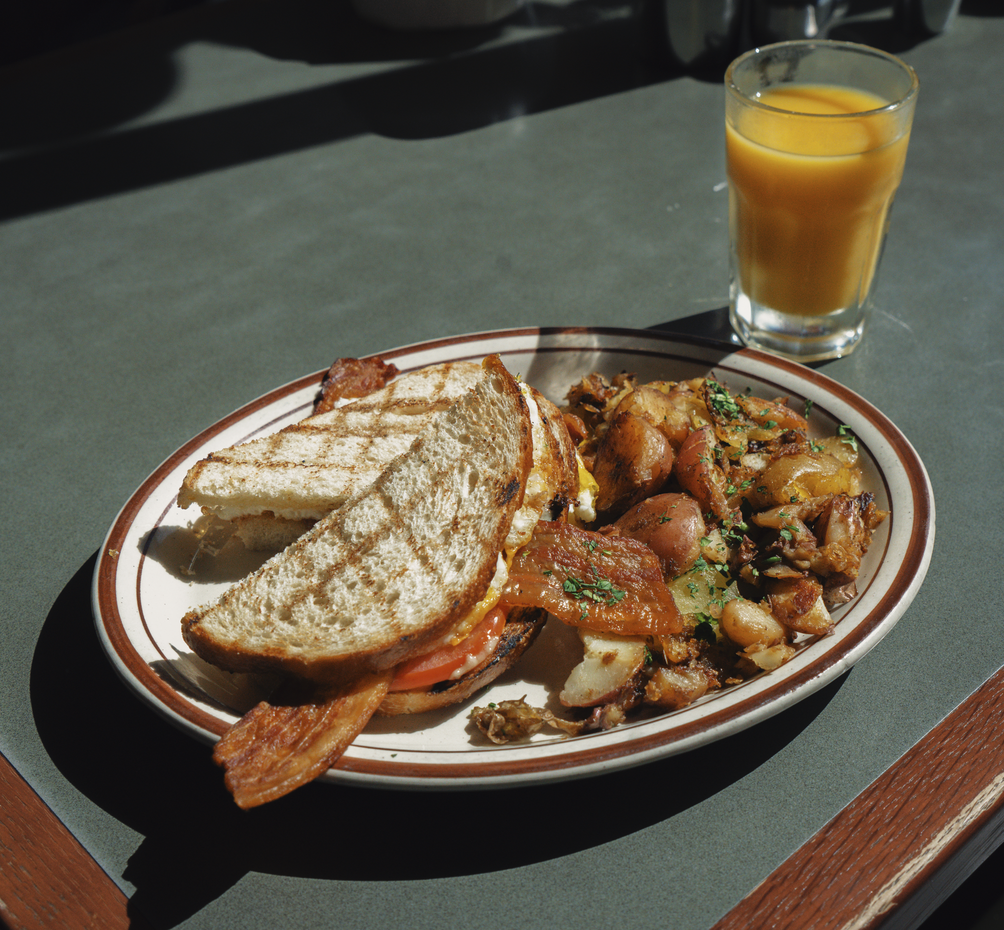 A halved Fried Egg Sandwich with bacon, served house potatoes and a glass of orange juice.
