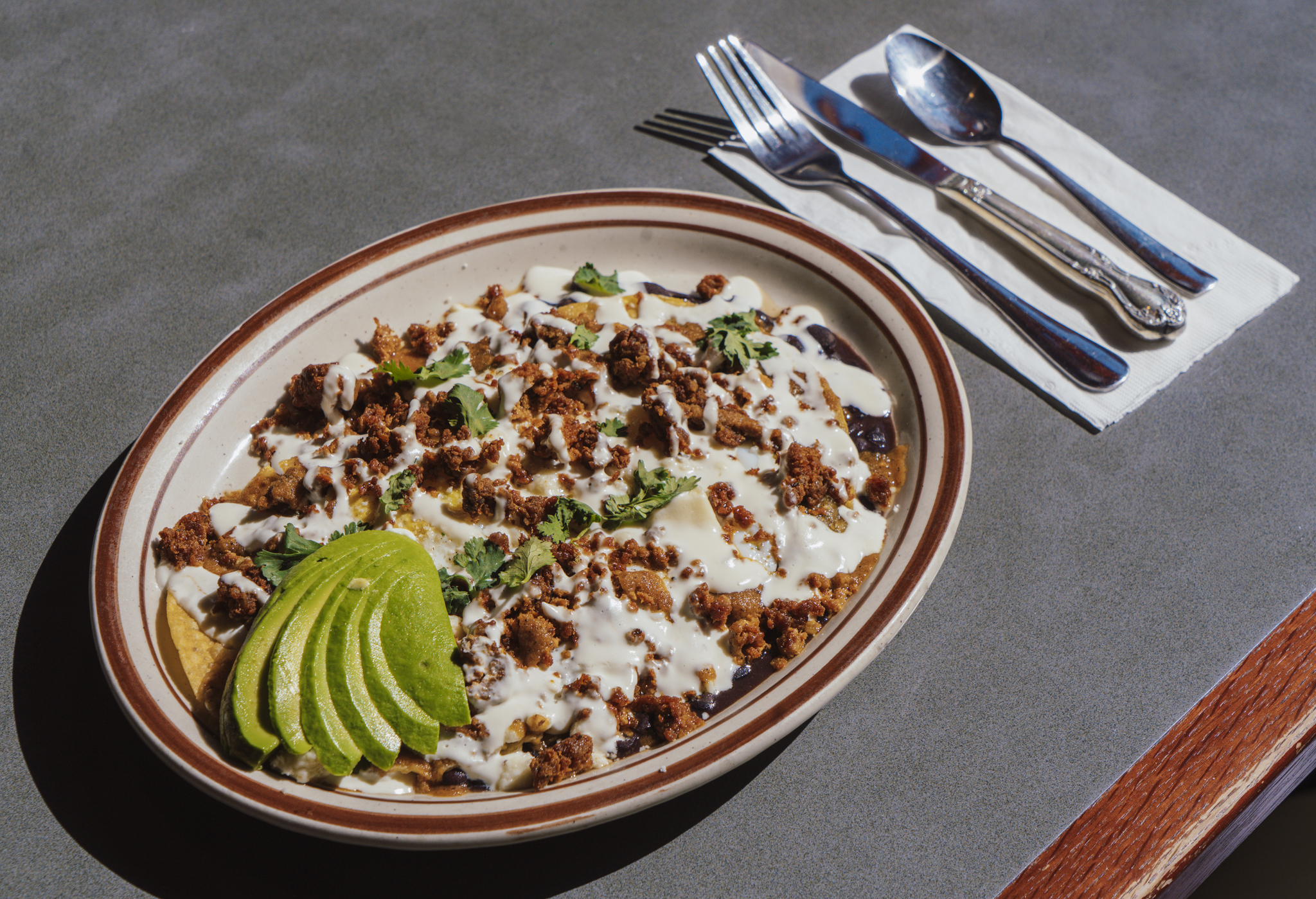 A plate of Huevos Rancheros topped with sliced avocado, presented next to a napkin and cutlery.