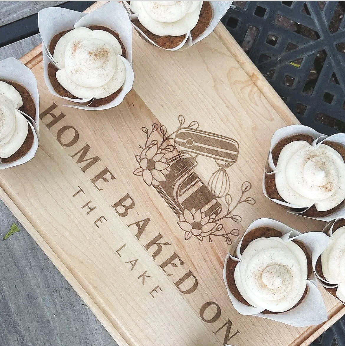 A wooden tray with the words "Home Baked on the Lake" and a floral illustration, surrounded by six cupcakes with white frosting in paper cups.