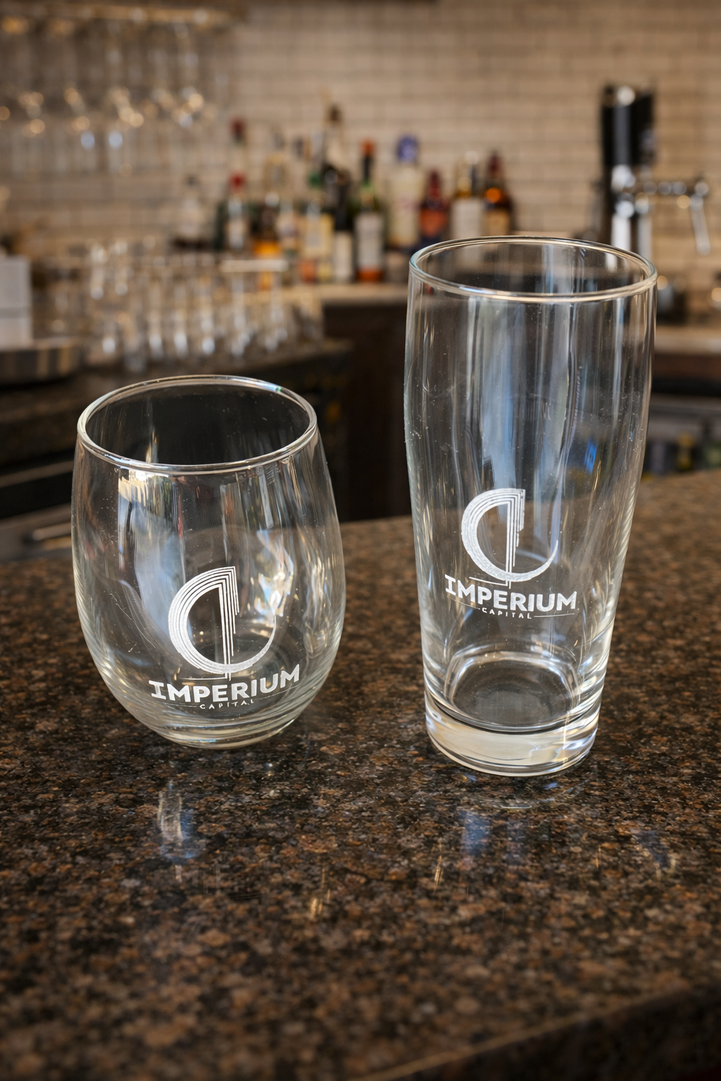 Two empty glass cups with 'Imperium Capital' logo on a dark granite bar counter, with a blurred background of a bar area with bottles and glasses.