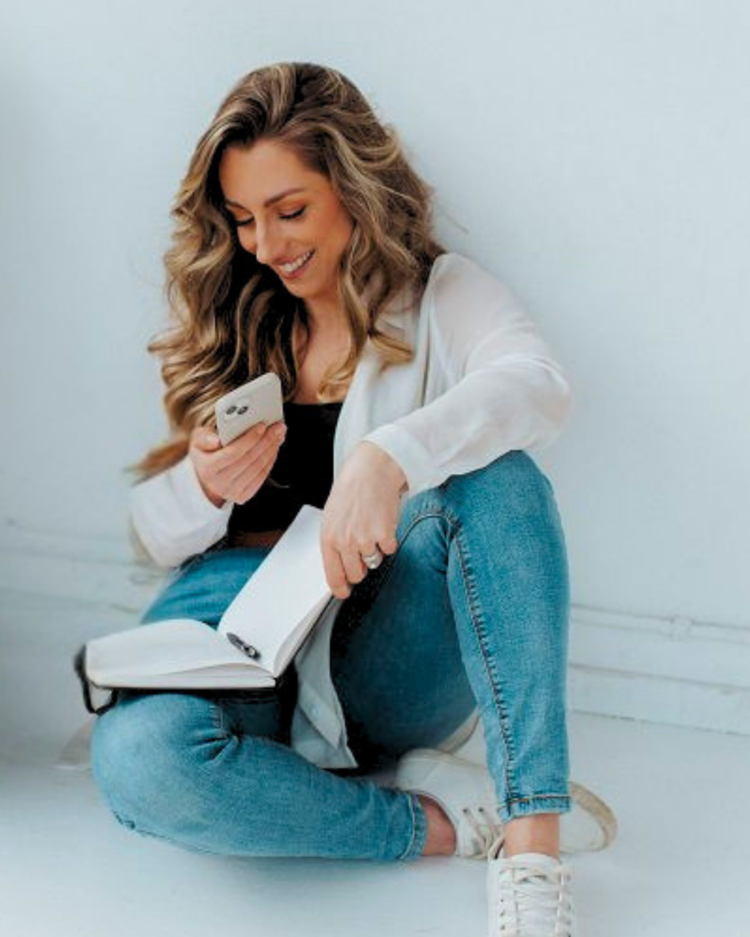A woman with long, wavy brown hair sitting on the floor, smiling, holding a smartphone and an open notebook with a pen, dressed in a white blazer, black top, blue jeans, and white sneakers, against a plain light-colored wall.