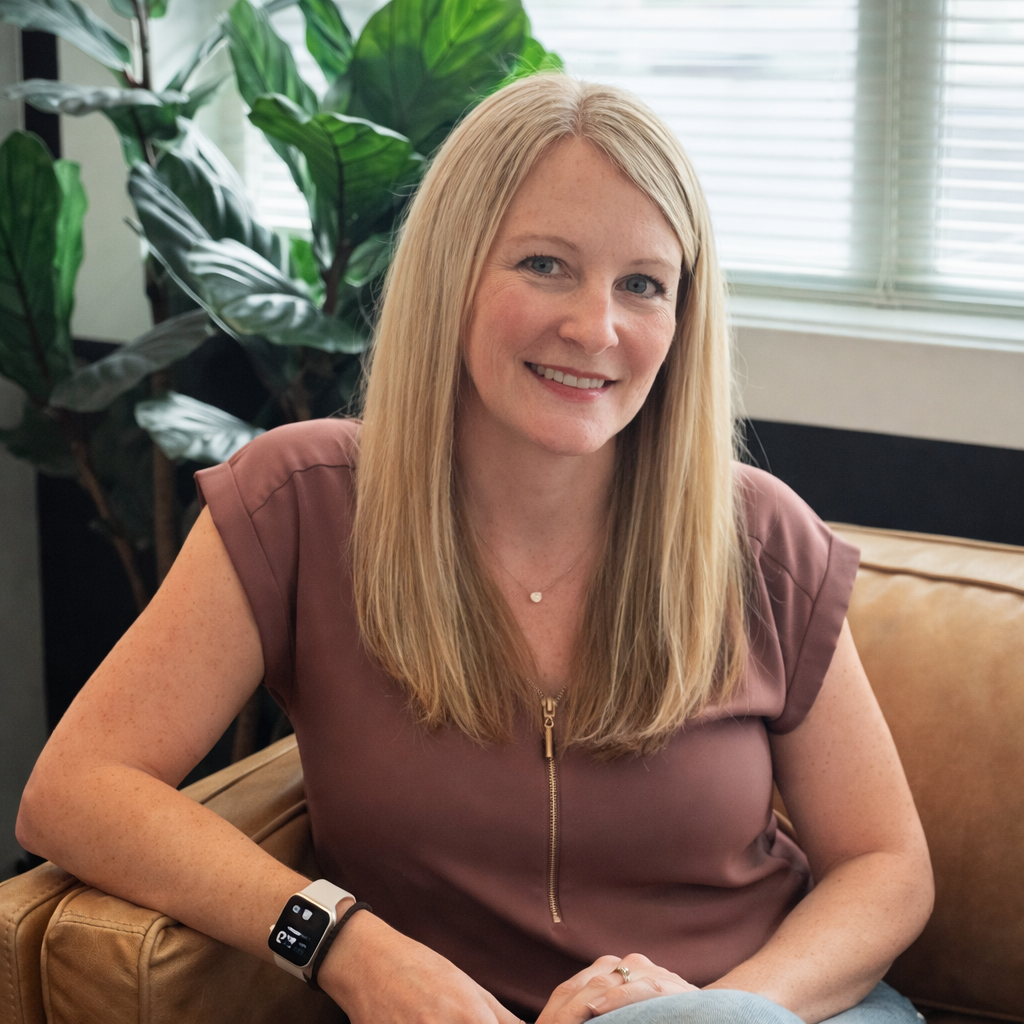 A young woman with long brown hair and fair skin standing outdoors in front of green foliage. She is smiling and wearing a navy blazer over a rust-colored top, with a delicate necklace.