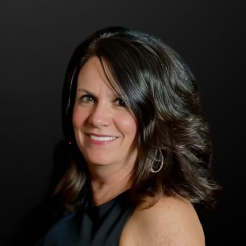 A woman with shoulder-length wavy dark brown hair with highlights, smiling, wearing a black top with a colorful abstract pattern, against a light neutral background.