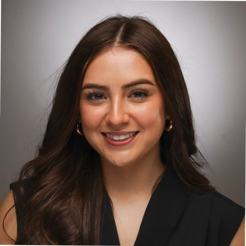 A woman with long dark hair smiling and crossing her arms outside in a park or garden area.