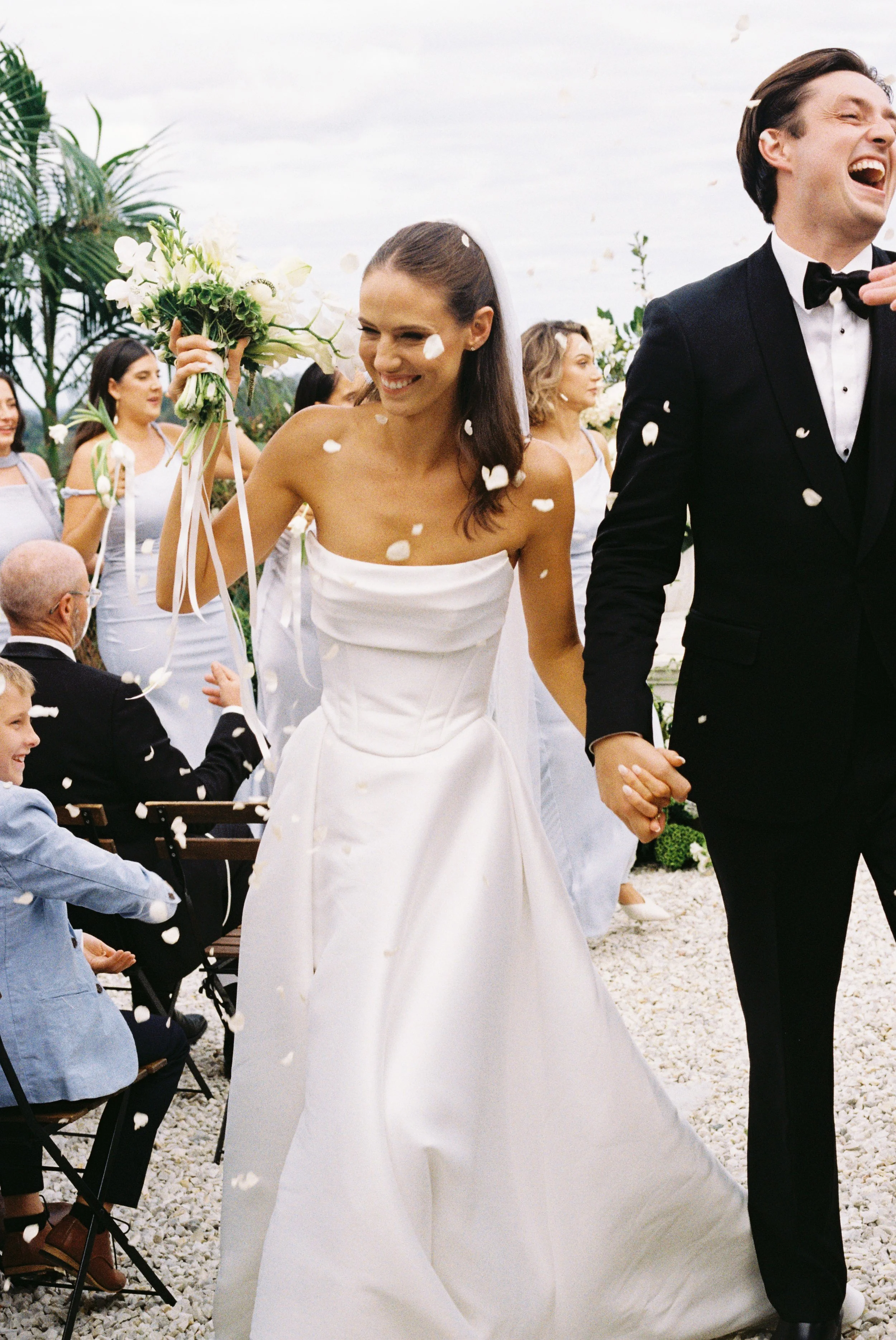 A bride and groom holding hands and smiling during their wedding celebration, surrounded by guests and flower petals in an outdoor setting.