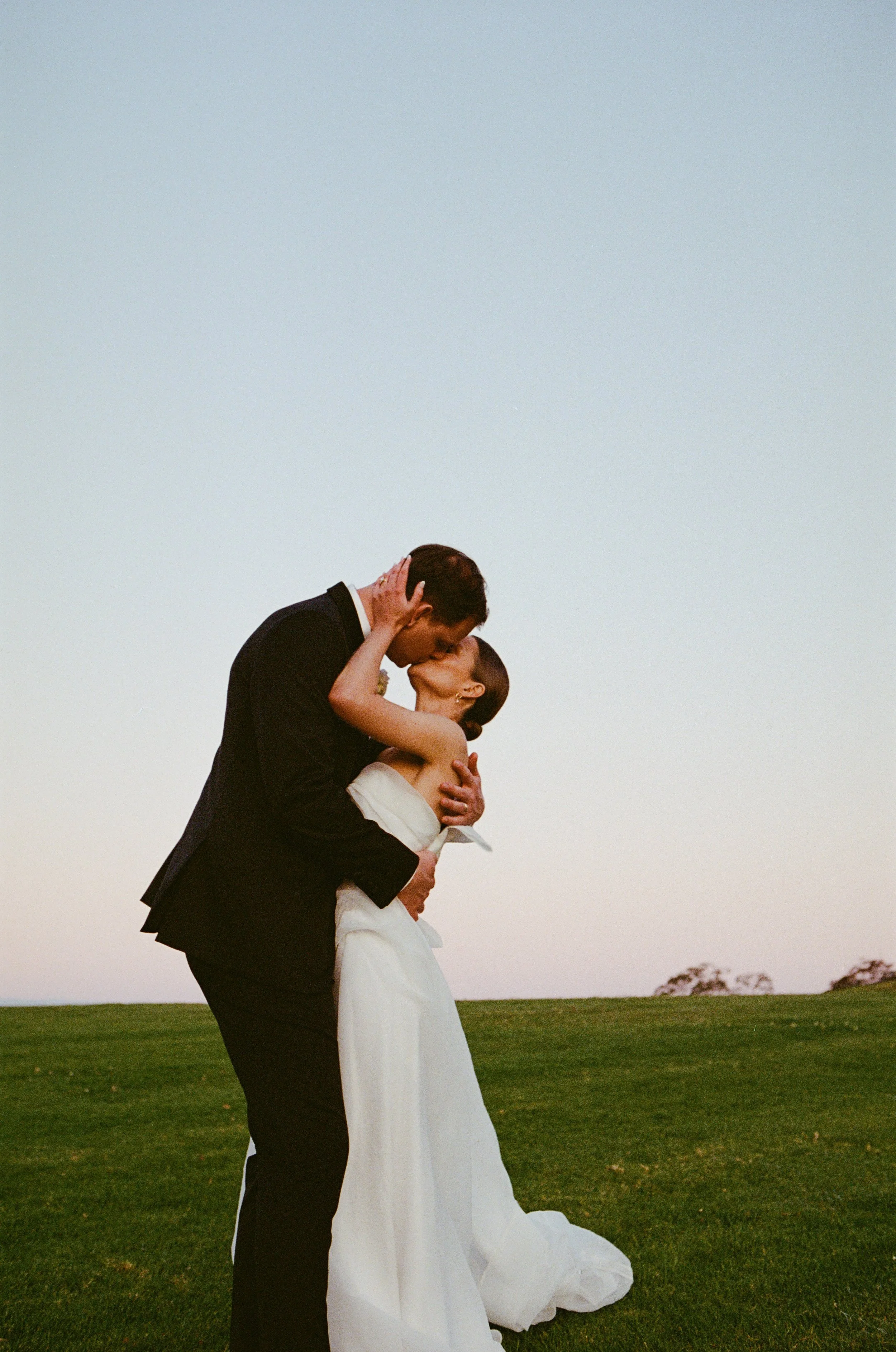 A couple is sharing a kiss outdoors on a grassy field during sunset, with the man in a black suit and the woman in a white wedding dress.