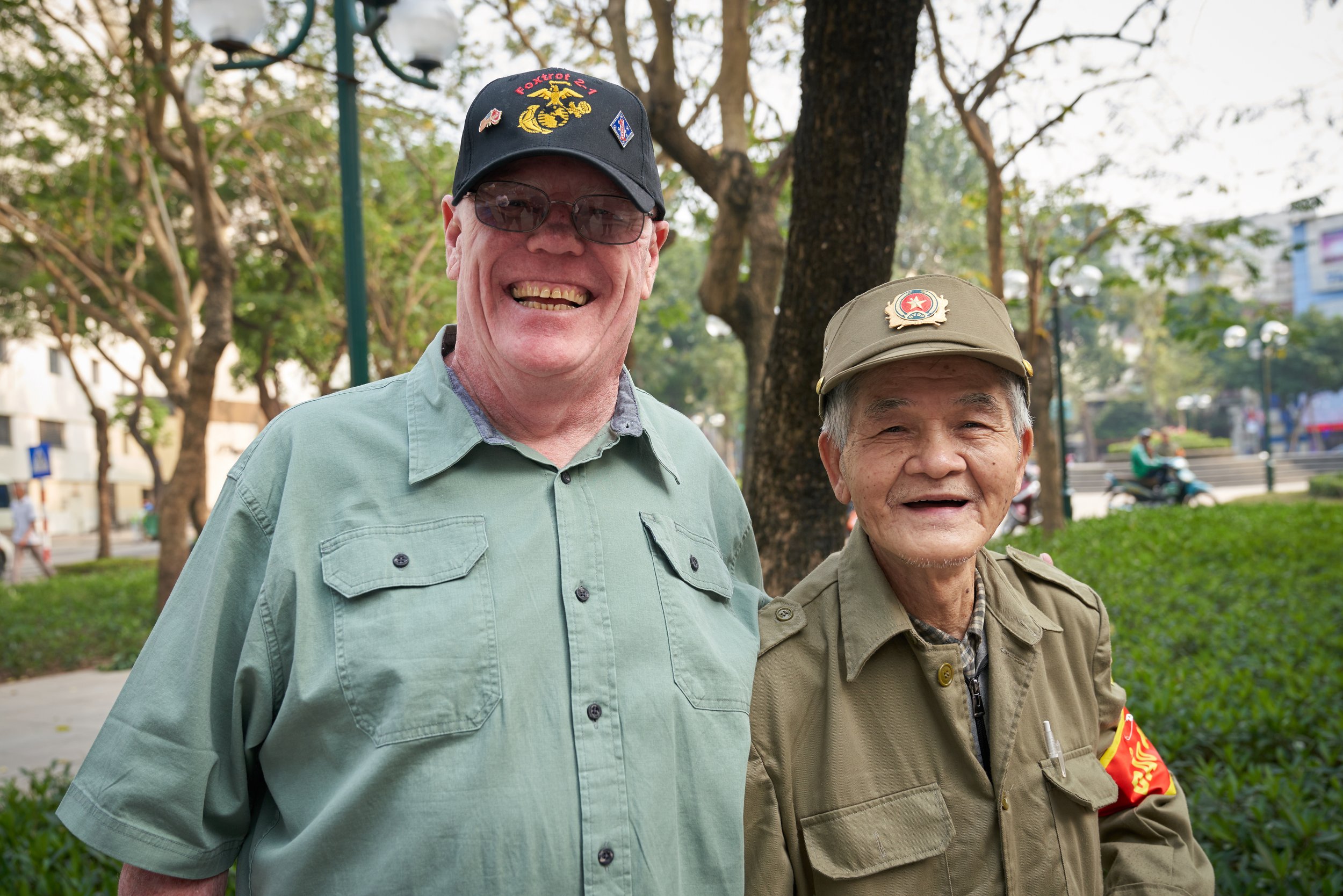 Two smiling men standing outdoors, one wearing a green shirt and black cap with the word 'Foxtrot' and the other in a green uniform with a matching cap, in a park setting.