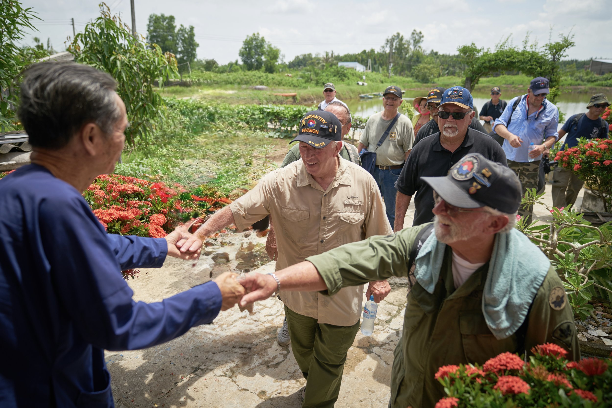 A group of men wearing military hats and casual clothing are walking outdoors in a rural area. One man is shaking hands with an elderly man in a blue traditional outfit. Red flowers are in the foreground, with green plants and a body of water in the