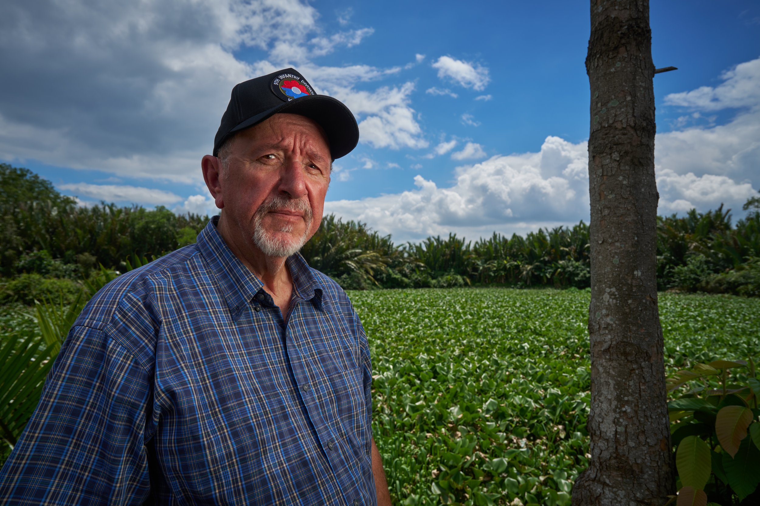 Man standing in a field with lush greenery and trees, wearing a plaid shirt and a cap, under a partly cloudy sky.