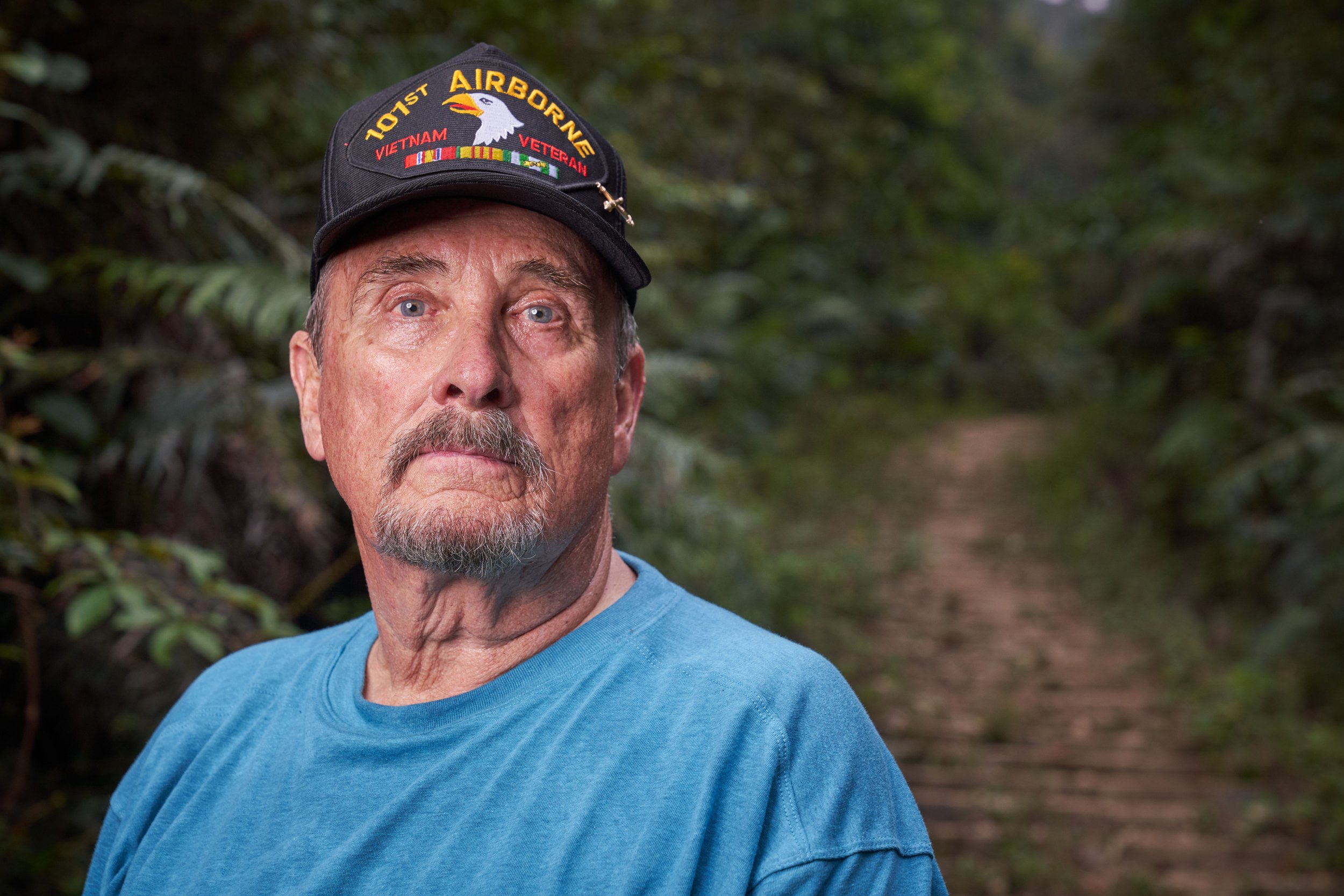 Portrait of an older man in a blue shirt wearing a military cap with '101st Airborne' and 'Vietnam Veteran' text, standing in a forest.