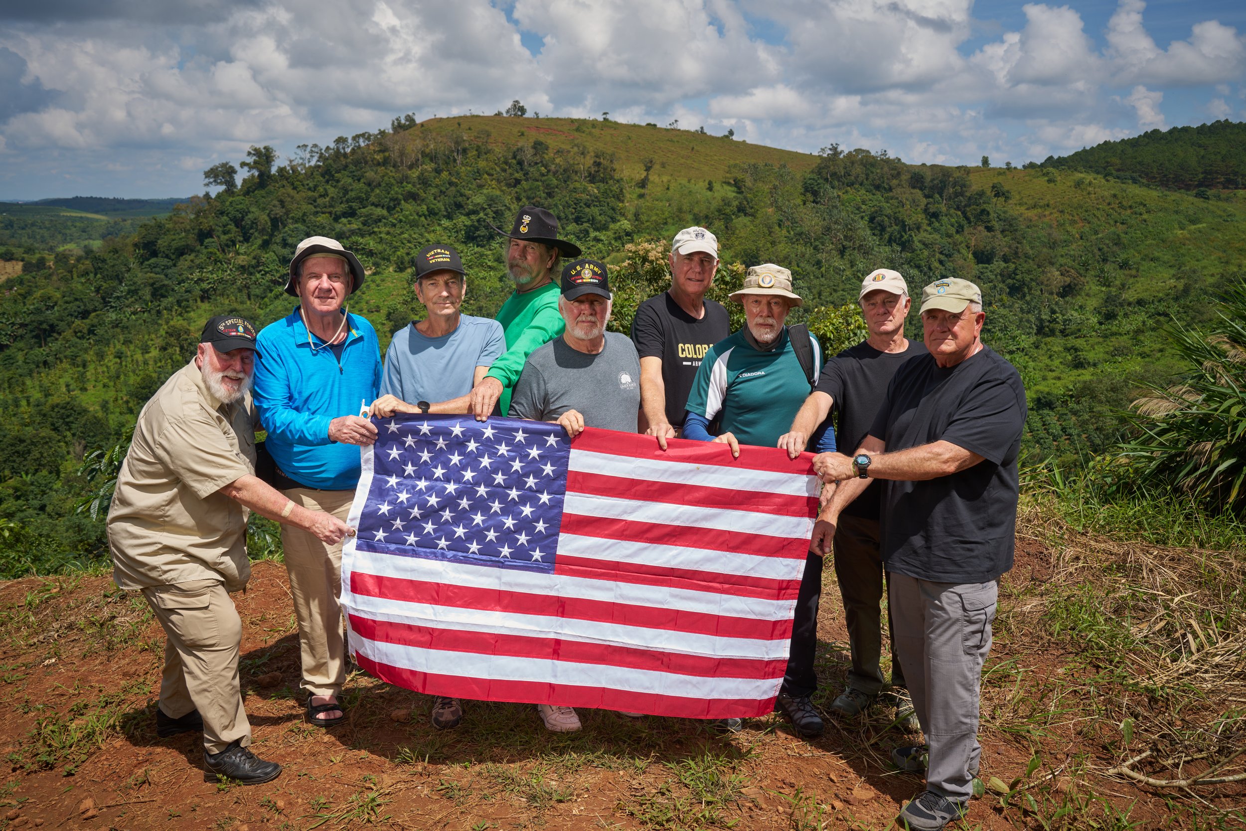 Group of men holding U.S. flag outdoors on grassy hillside with trees in background
