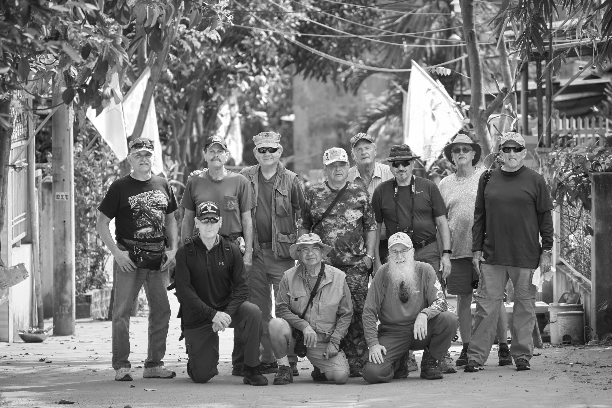 A group of eleven men posing outdoors in a garden or park, some standing and some kneeling, surrounded by trees and plants.