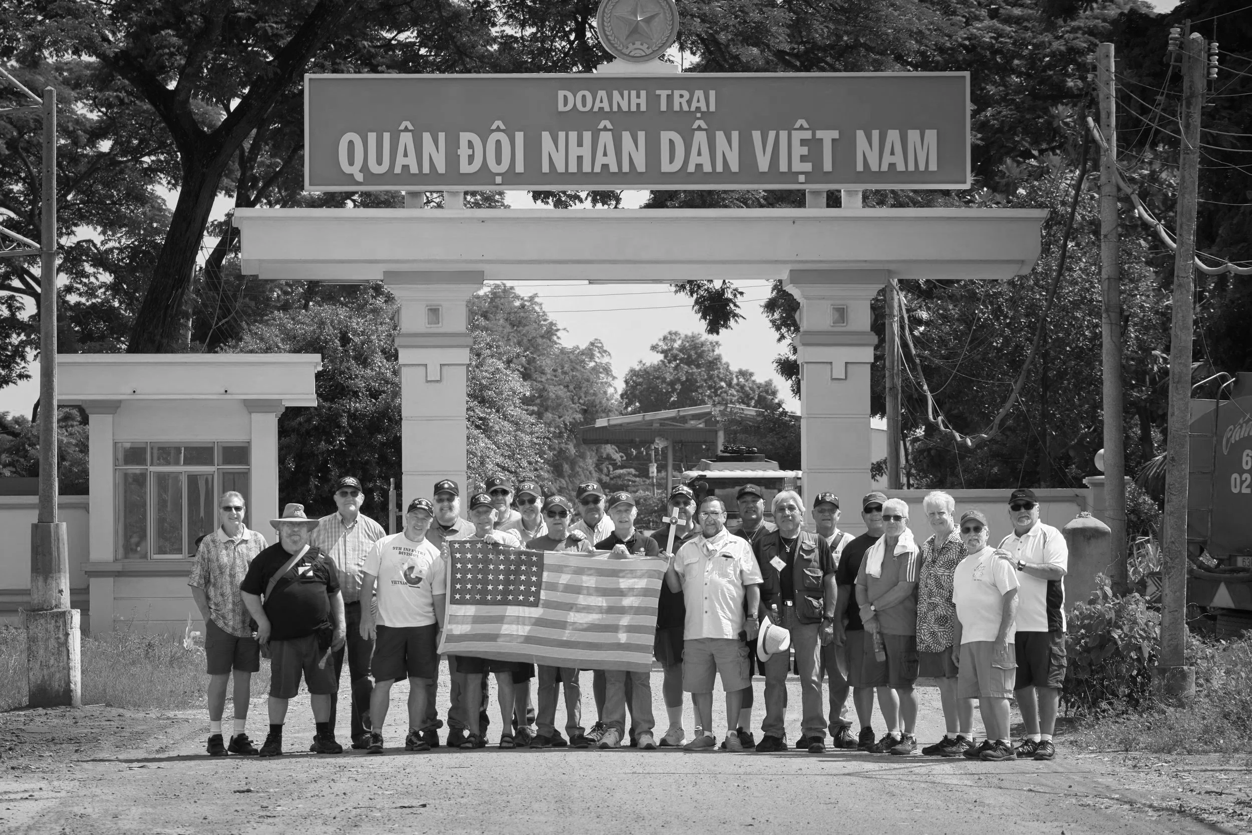 A group of people holding an American flag standing under a sign in Vietnamese script, with trees and utility poles in the background.