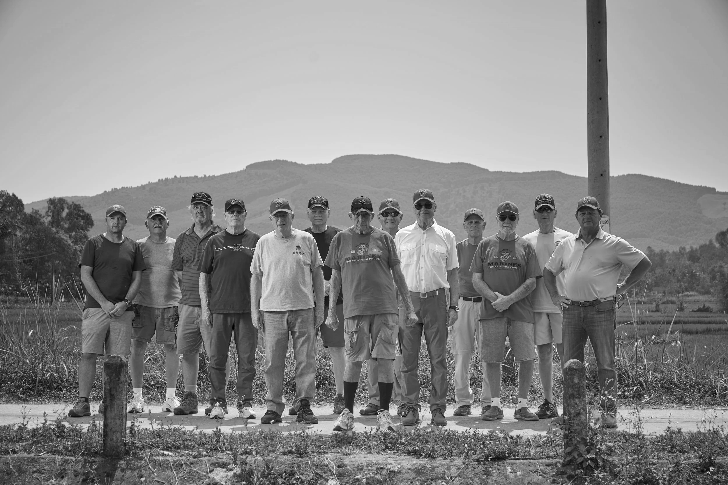 Group of 13 men standing outdoors on a road with green fields and mountains in the background, some wearing baseball caps and casual clothing.