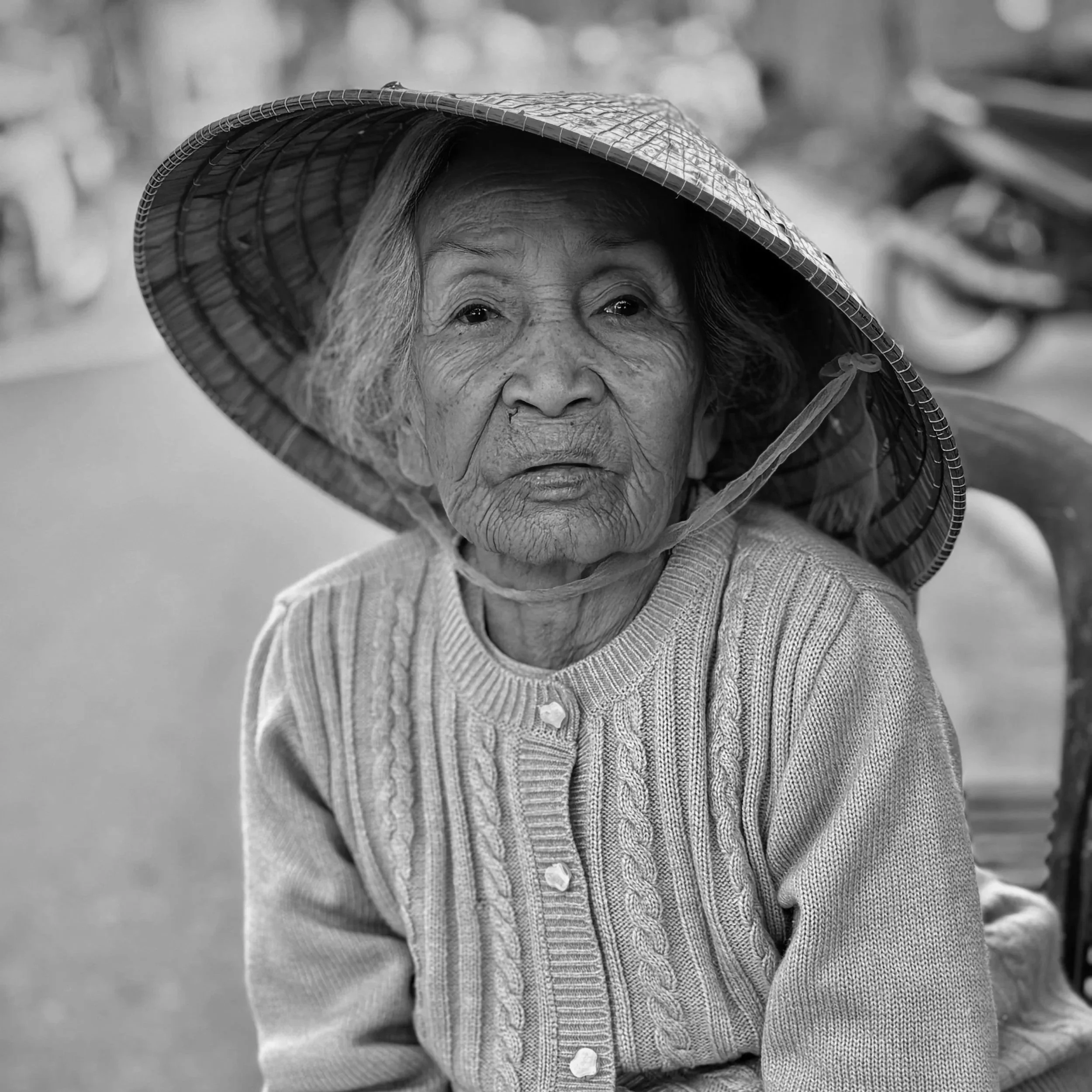 Black and white photo of an elderly Asian woman wearing a wide-brimmed straw hat and a knitted sweater, sitting outdoors.