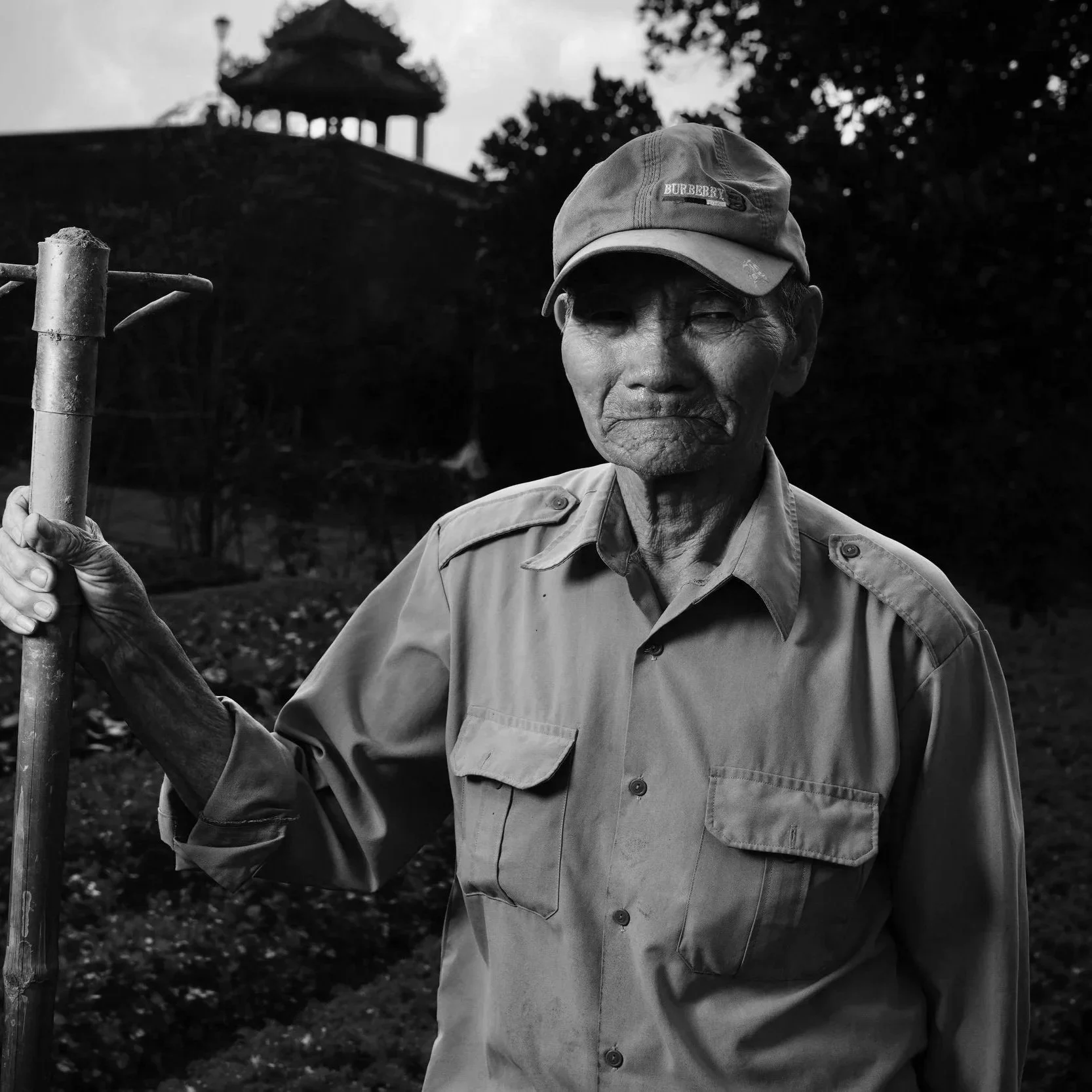 A black and white photo of an elderly man wearing a baseball cap and a button-up shirt, standing outdoors with trees and a pavilion in the background. He is holding a gardening tool.
