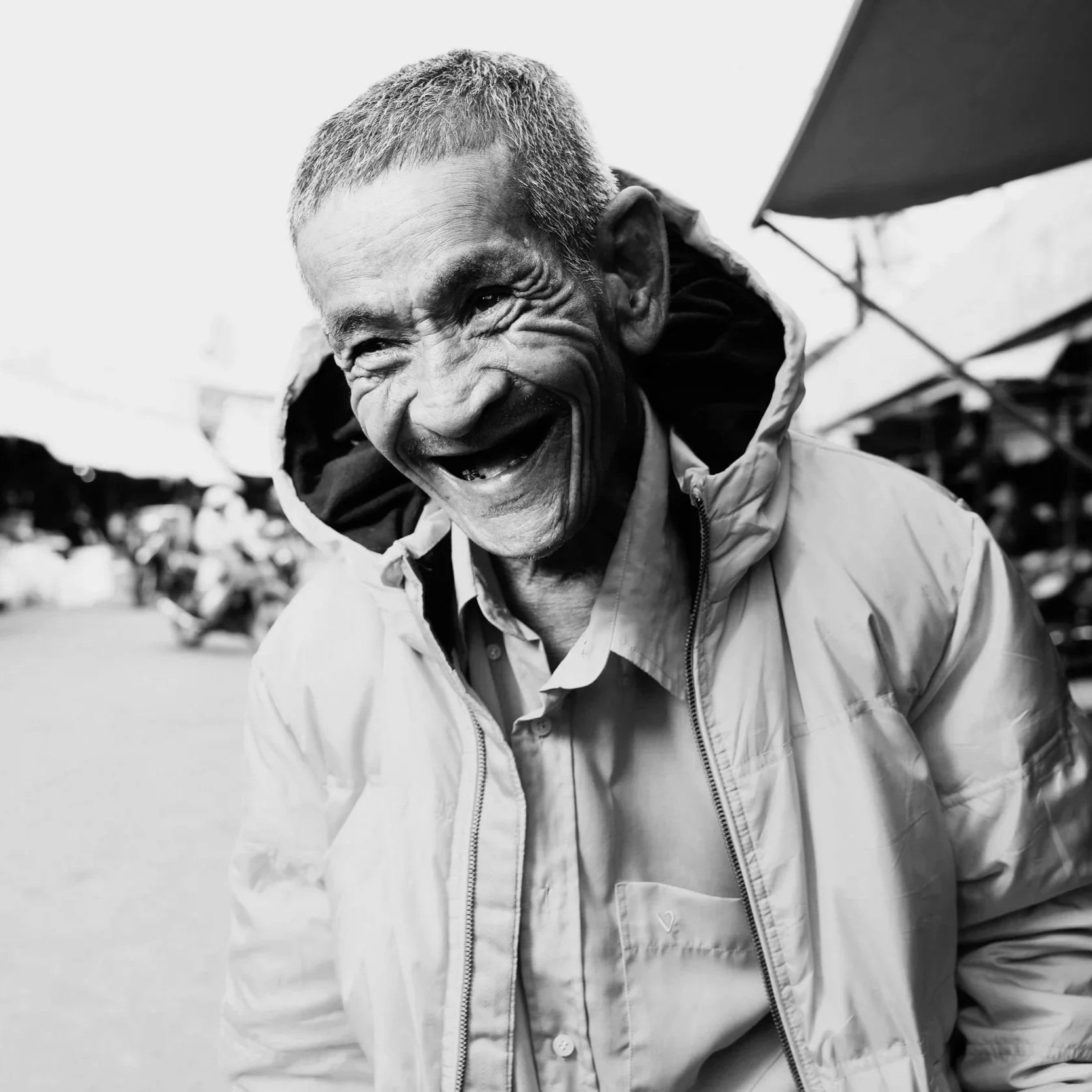 Black and white photo of an elderly man smiling and looking joyful, wearing a jacket, outdoors with market stalls in the background.