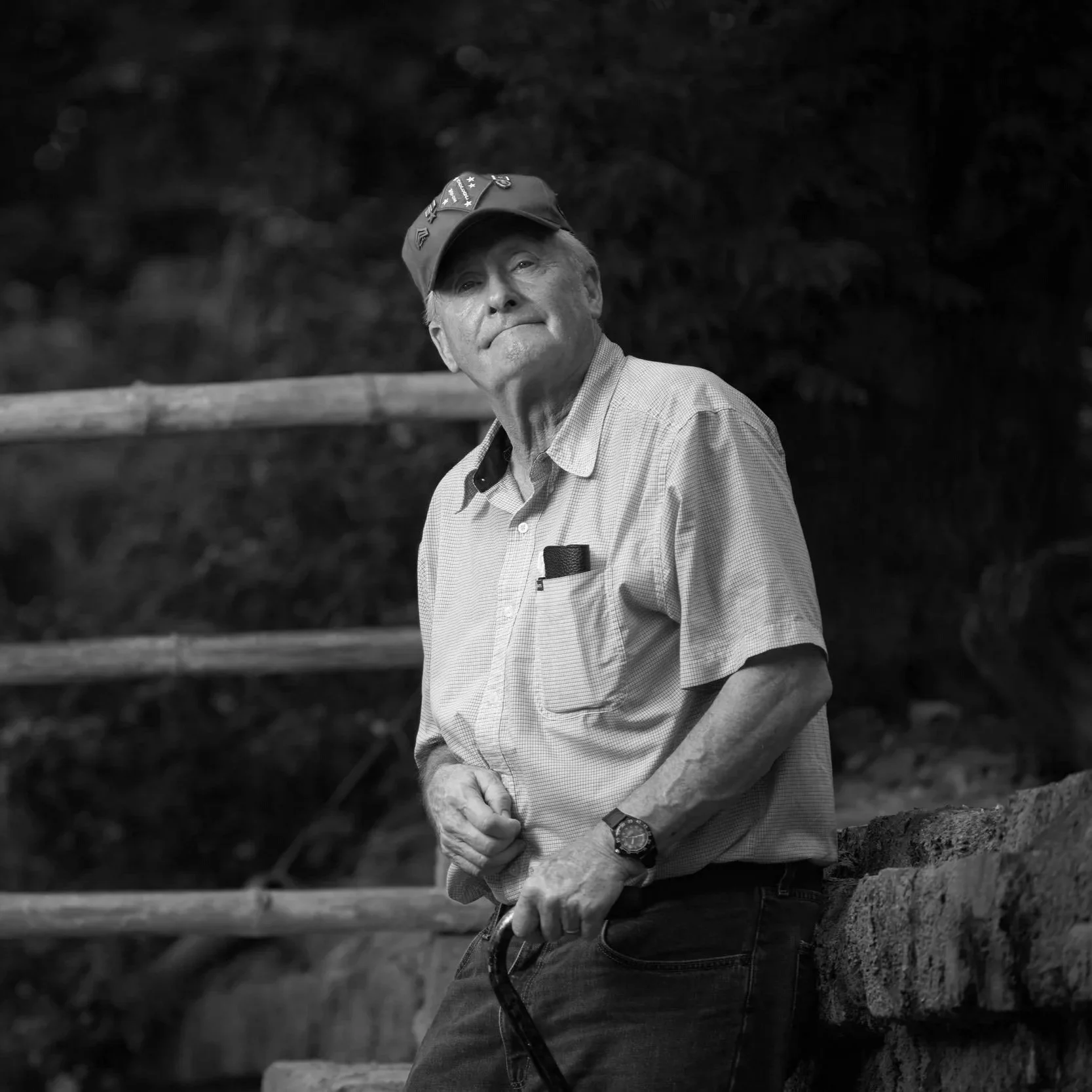 An elderly man with a cap, striped shirt, and a wristwatch, standing outdoors near a stone wall and wooden railing, looking to the side.
