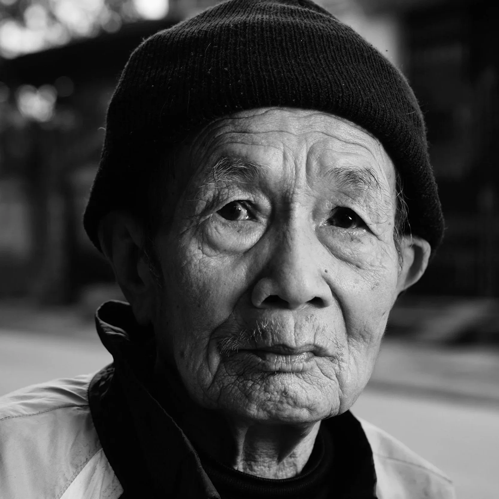 Close-up black and white portrait of an elderly Asian woman wearing a beanie and a scarf, with a calm expression.