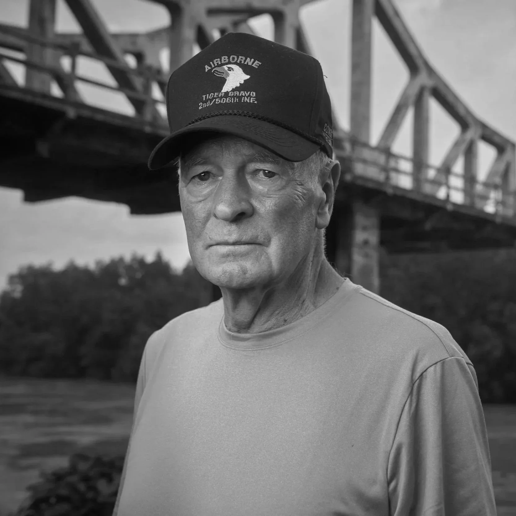 A man with a serious expression wearing a cap that says 'Airborne' and 'Tiger Bravo', standing outdoors near a bridge.