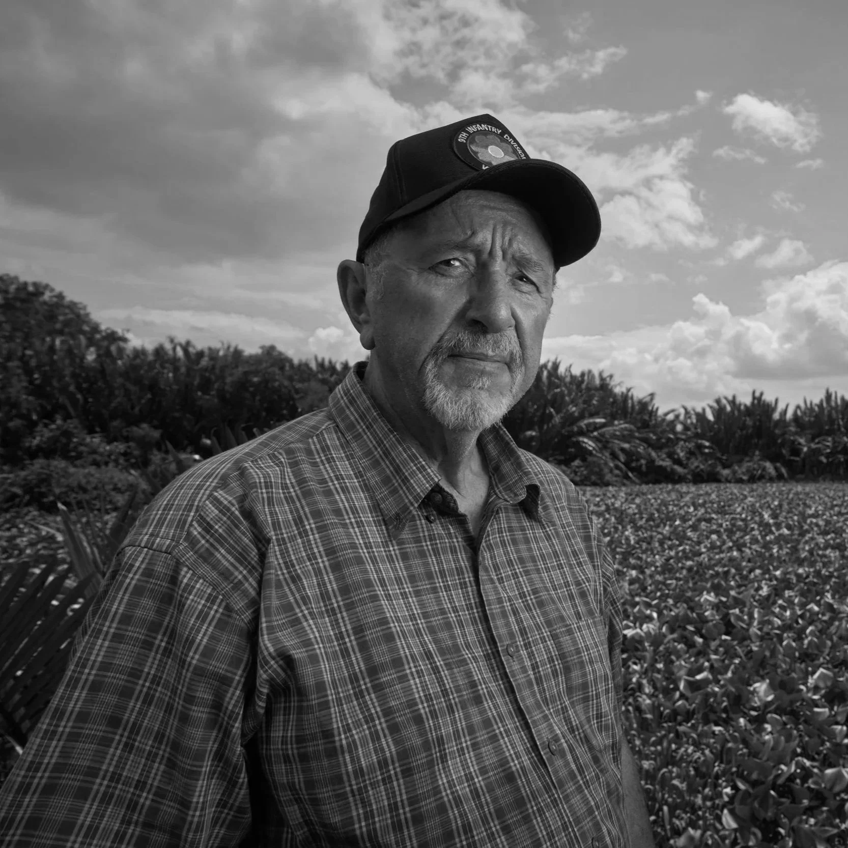 A black-and-white photo of an older man with a beard, wearing a baseball cap and a plaid shirt, standing outdoors with a field of plants and bushes behind him, under a partly cloudy sky.