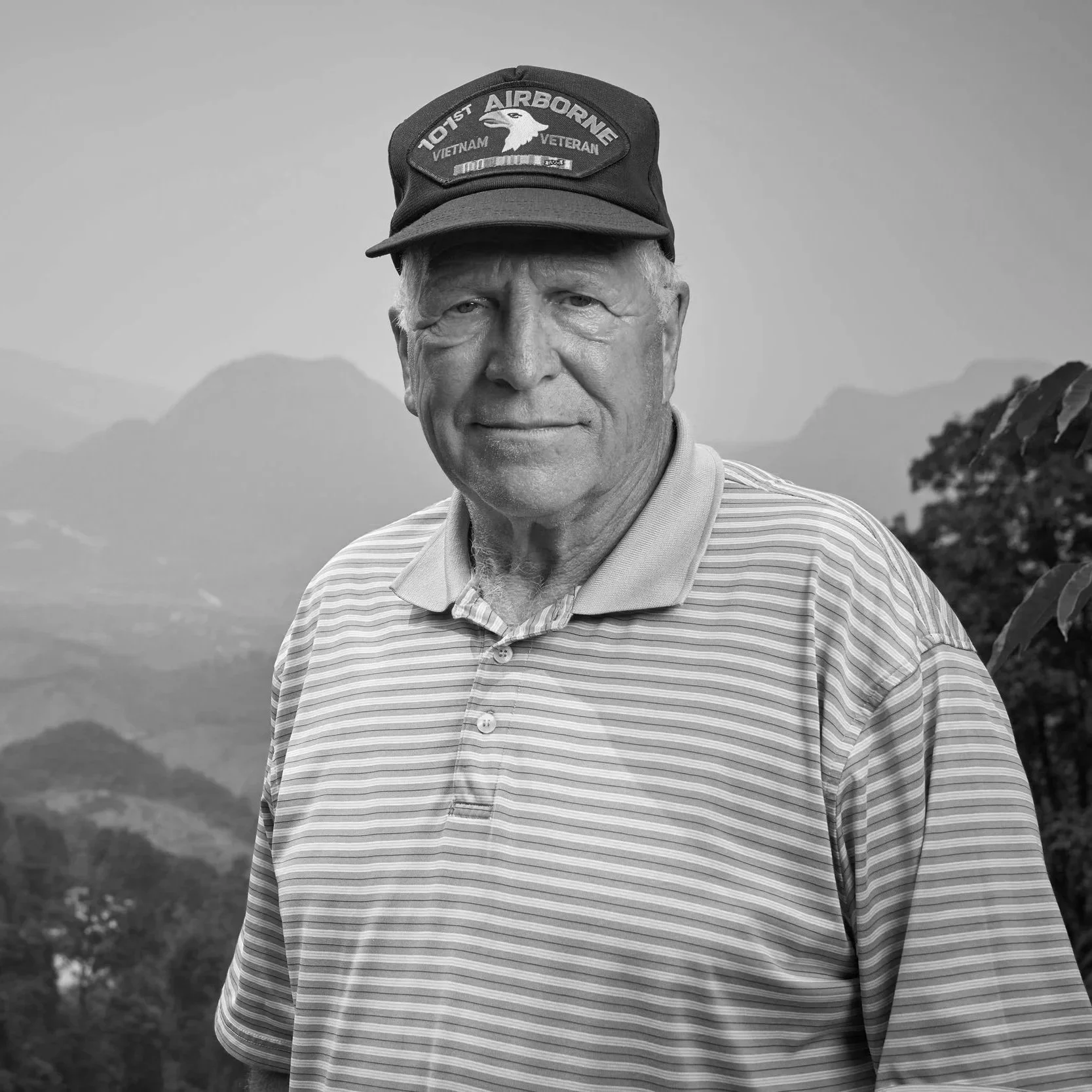 A black and white photo of an elderly man wearing a cap that reads 'VTOIST AIRBORNE VIETNAM VETERAN' and a striped polo shirt, with a mountainous landscape in the background.