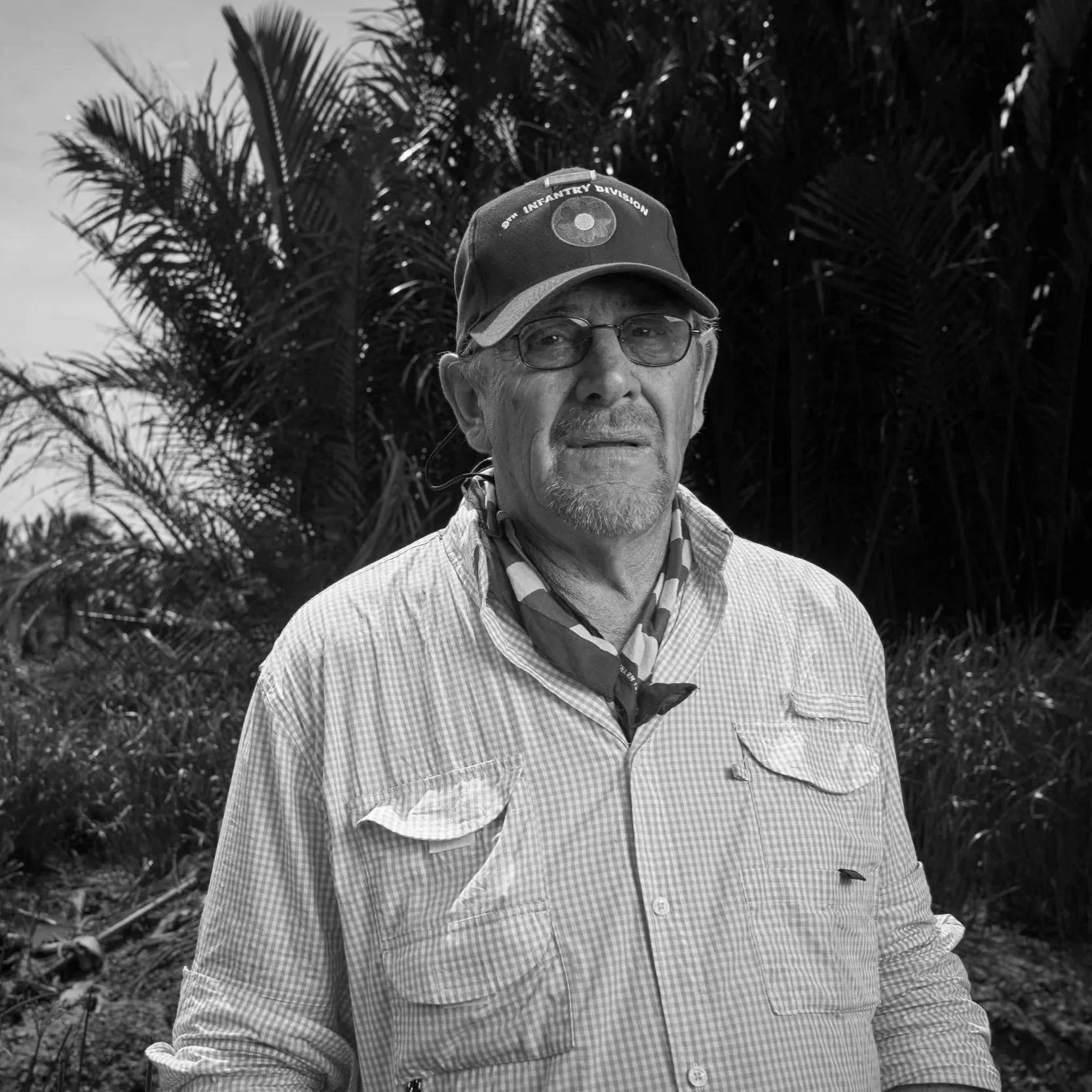 An elderly man wearing glasses, a cap with a patch, a checkered shirt, and a bandana around his neck, standing outdoors with palm trees in the background.