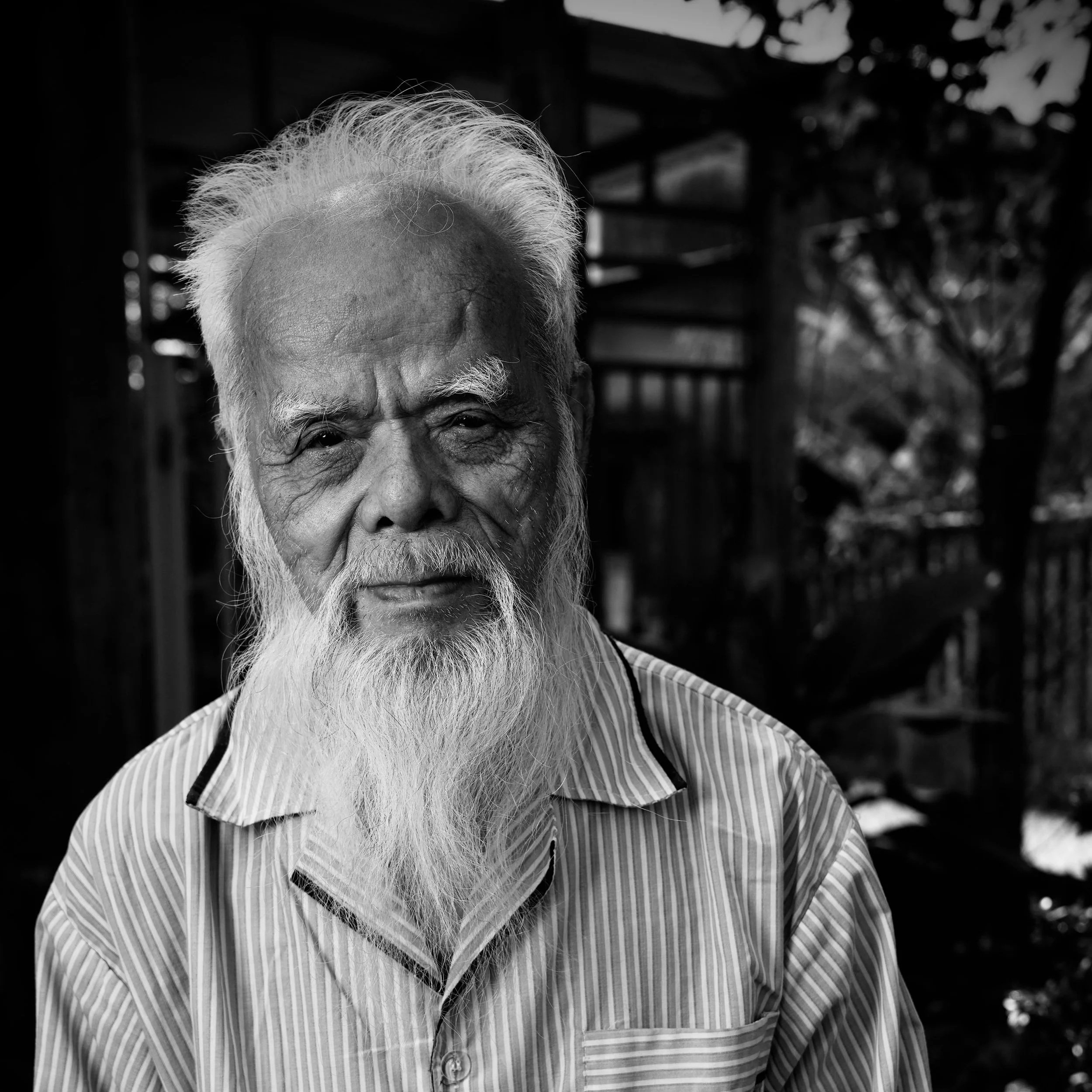 A black and white photo of an elderly man with a long white beard and hair, wearing a striped shirt, outdoors with trees and wooden structures in the background.