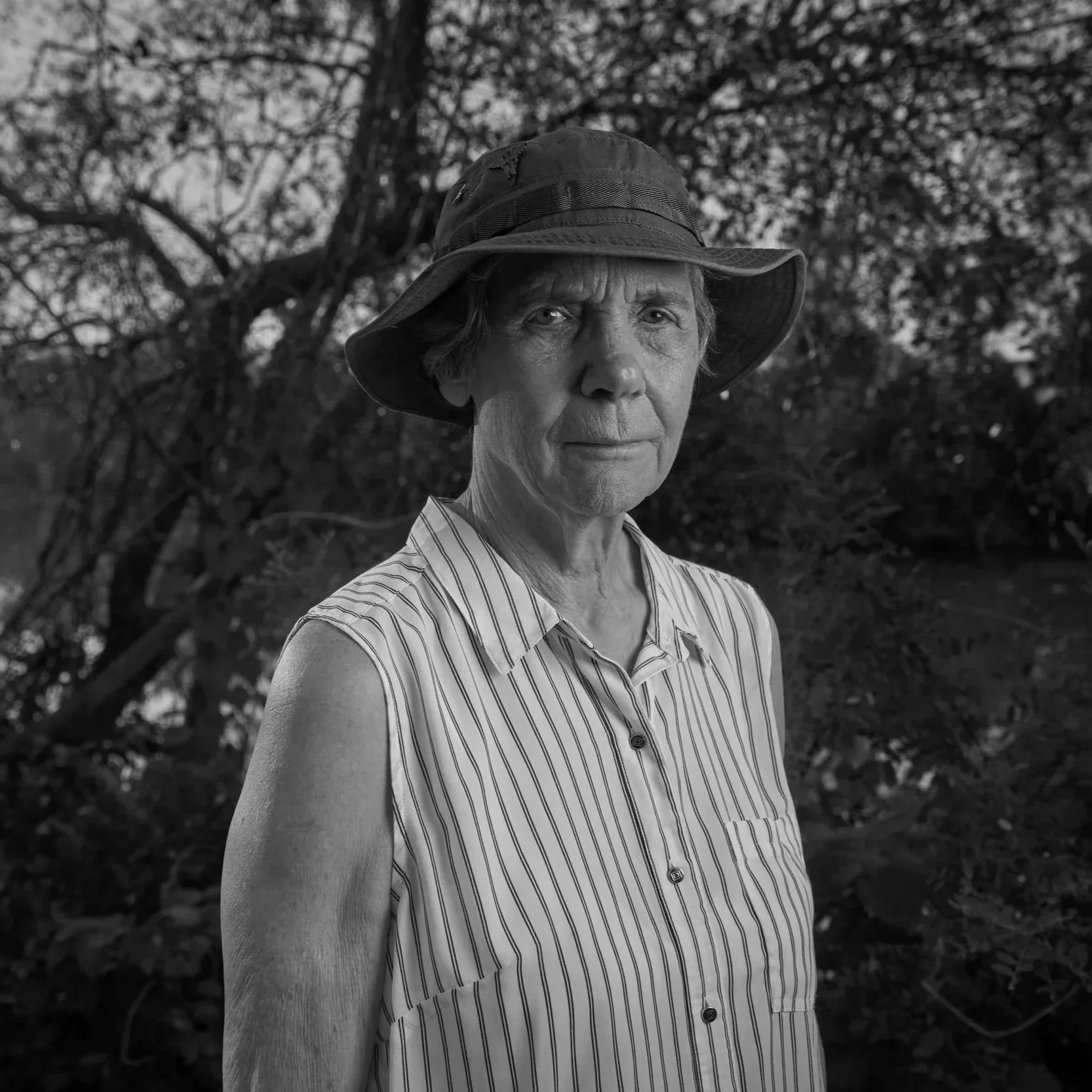 A black and white portrait of an elderly woman wearing a striped sleeveless shirt and a wide-brimmed hat, standing outdoors with trees in the background.