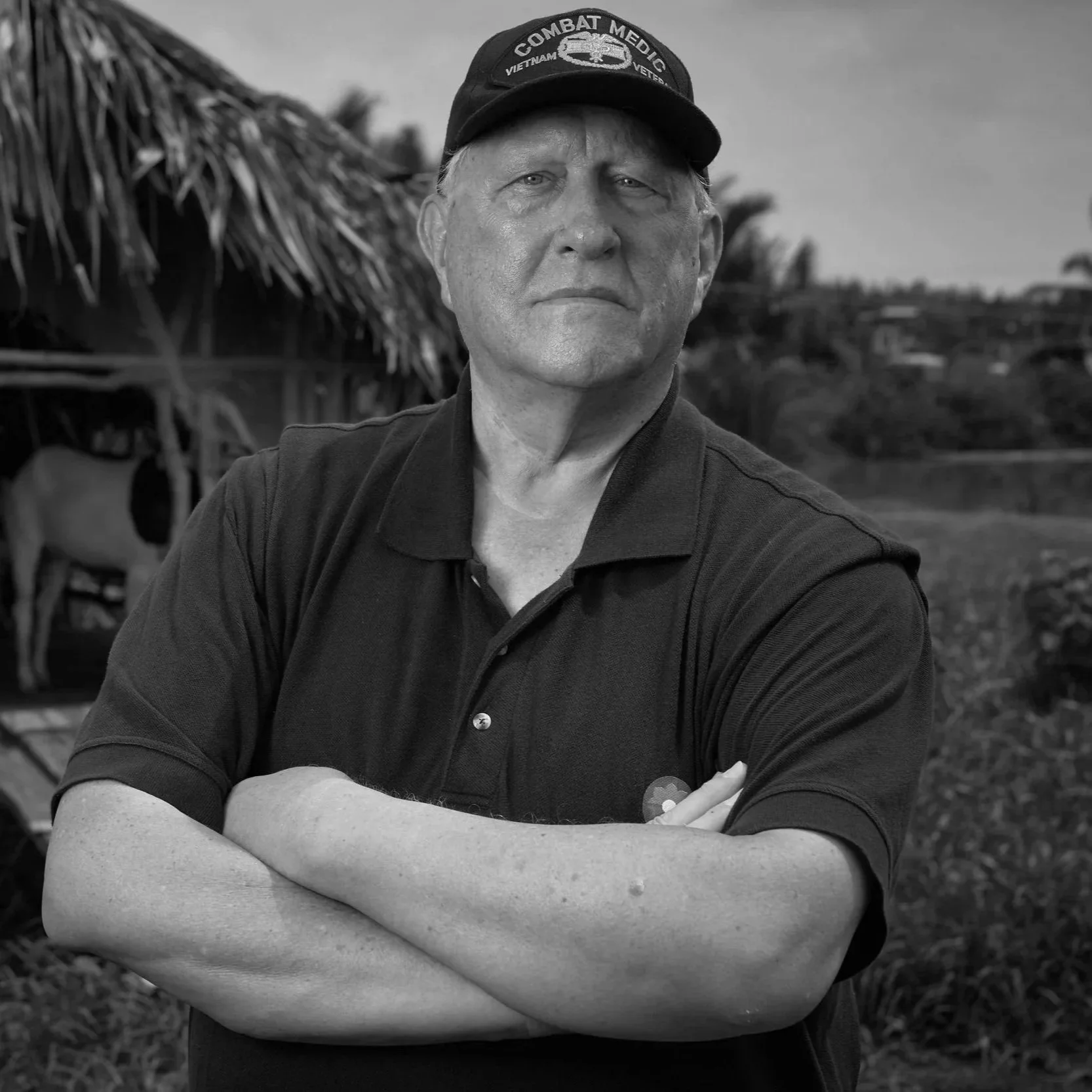 A man in a black polo shirt with folded arms is standing outdoors in front of a thatched-roof structure and a wooden vehicle, wearing a military-style cap that says 'Combat Medic.'