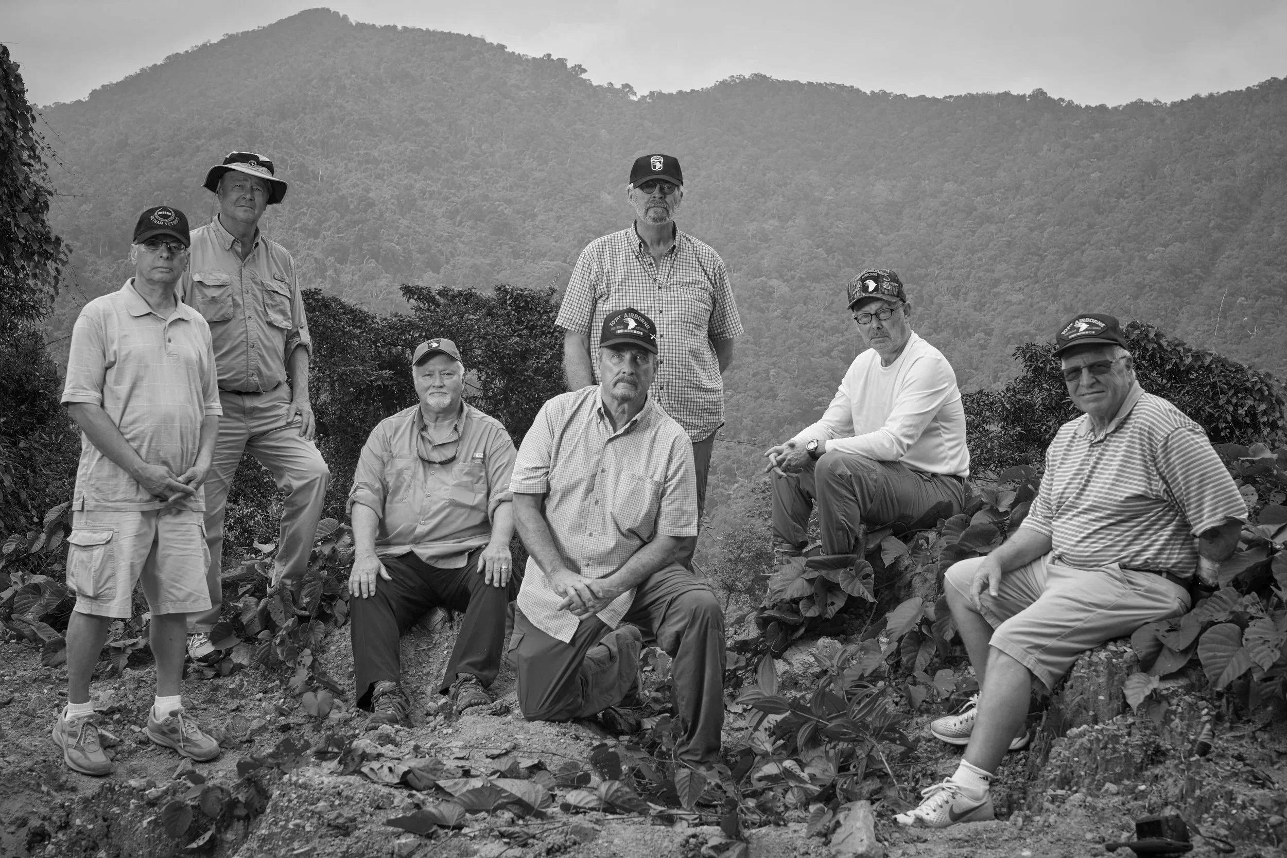 Black and white photo of eight men outdoors on a hillside with mountains in the background. They are dressed in casual outdoor clothing, some wearing hats and sunglasses, and are positioned among plants and soil.
