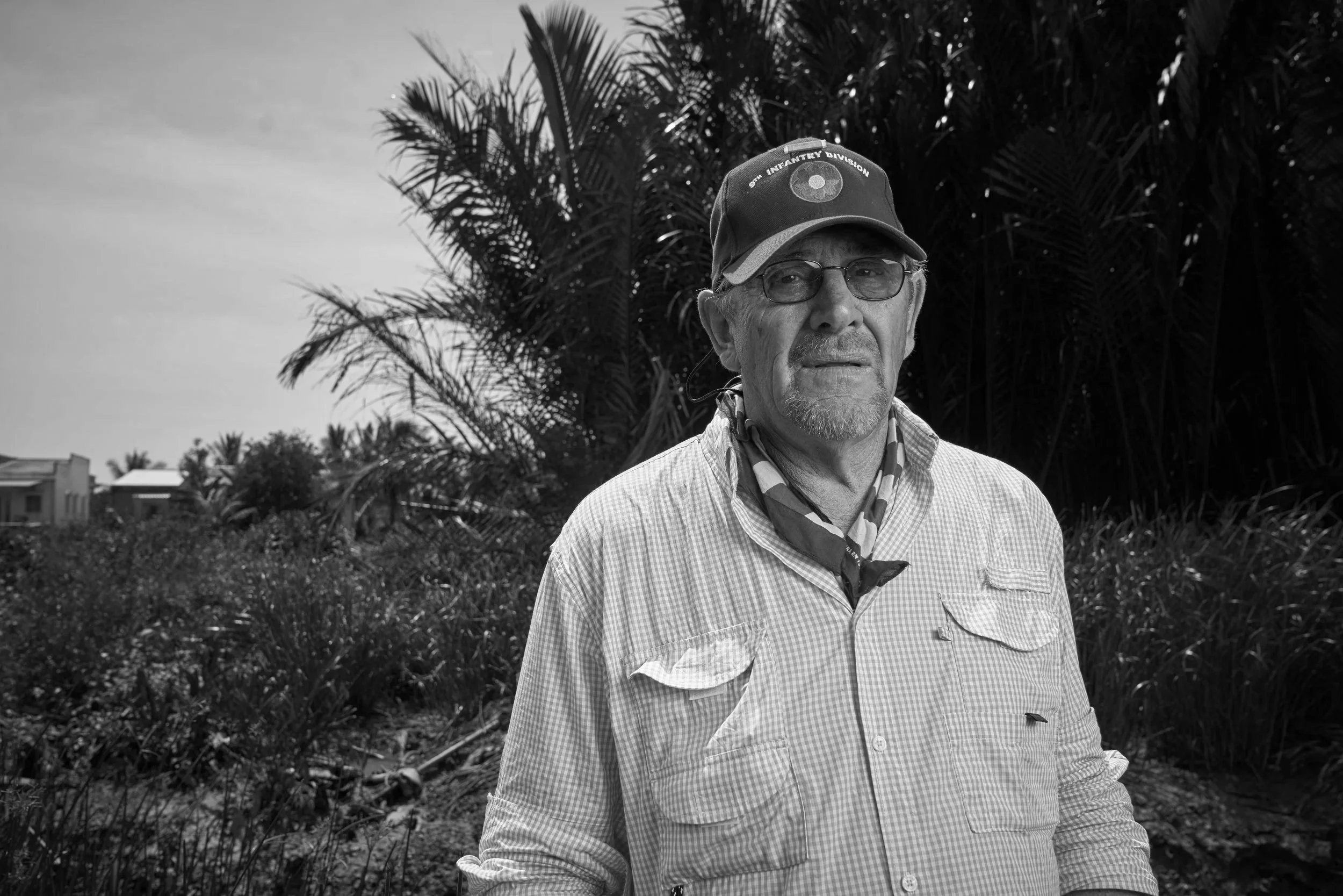 An elderly man wearing a navy cap with a patch that reads 'Infantry Division', glasses, a blue and white checkered shirt, and an American flag patterned bandana around his neck standing outdoors in a lush green area with palm trees and cloudy sky.