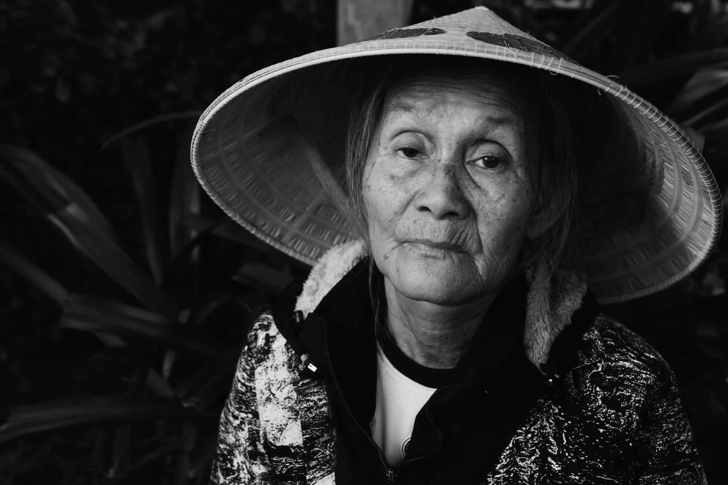 An elderly woman wearing a large traditional Asian conical hat outdoors, surrounded by green plants.