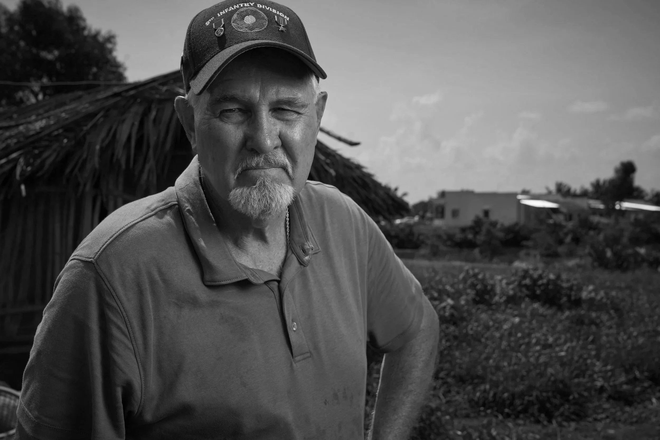 An older man with a white beard and mustache, wearing a beige polo shirt and a blue baseball cap with military badges, standing outdoors in a rural setting with green fields and a thatched roof structure in the background.