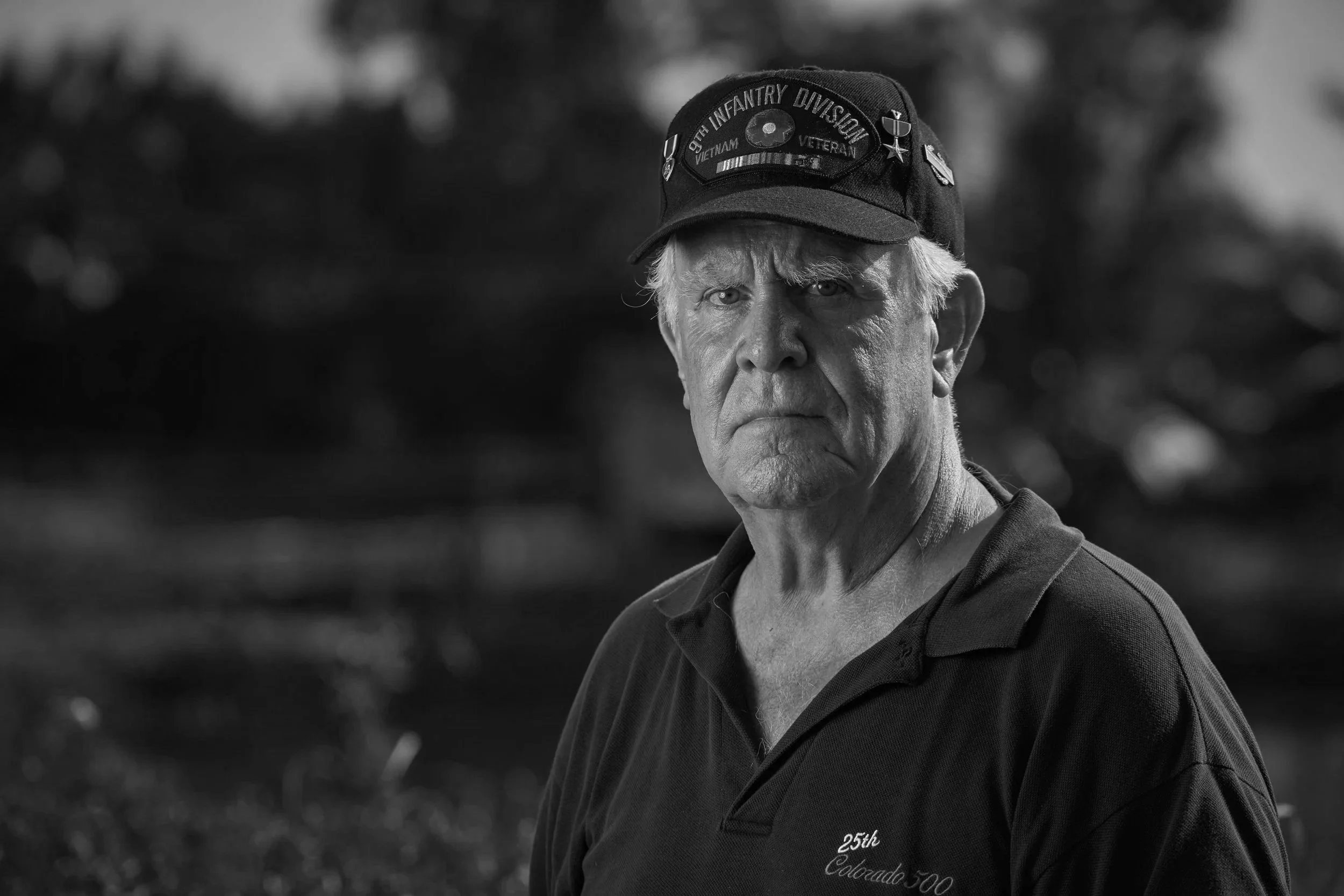 An elderly man with gray hair and a serious expression, wearing a black cap with various military and veteran patches, outdoors with a green, blurred background.