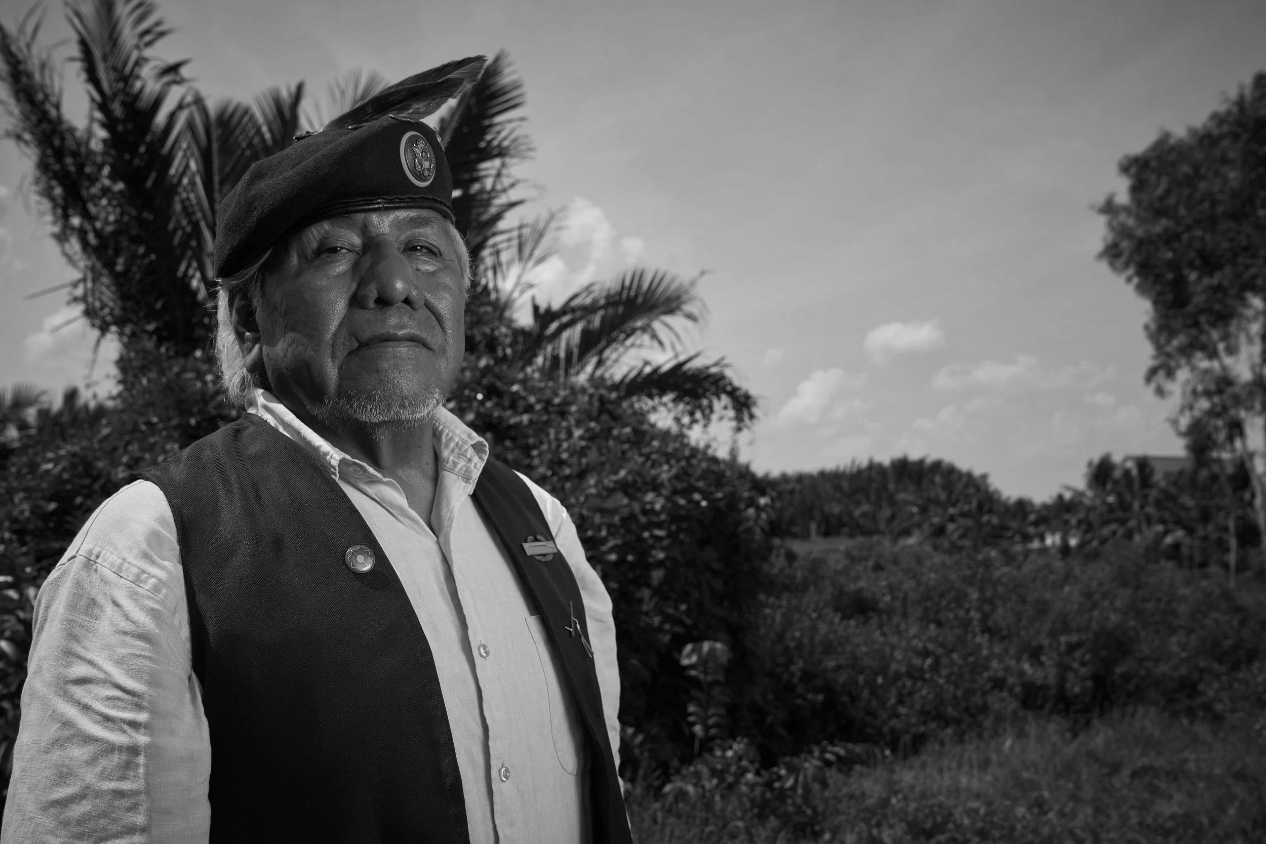 An elderly indigenous man wearing a black feathered headdress, white collared shirt, and black vest stands outdoors amidst greenery, with a blue sky and clouds in the background.