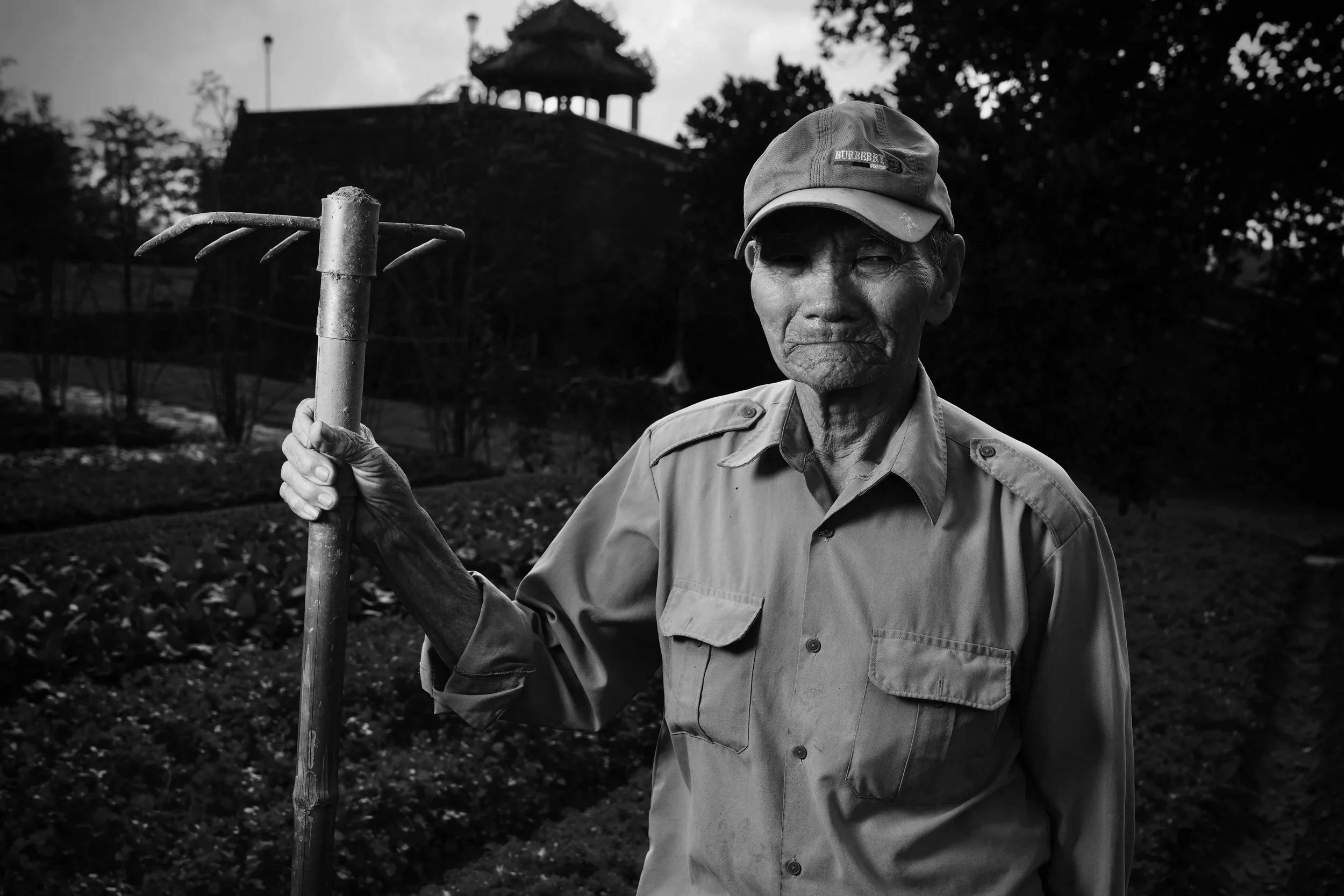 An elderly man holding a farming tool in a green field with trees and a wooden structure in the background.