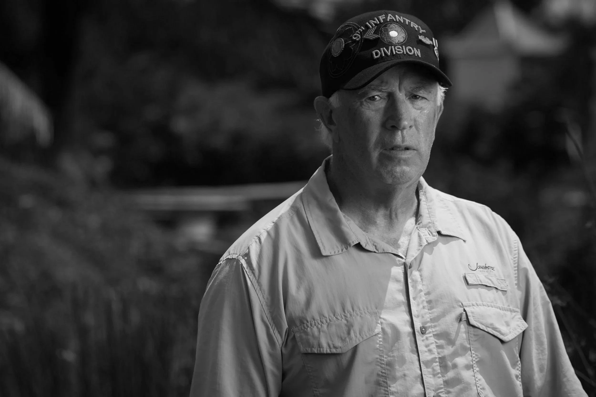 A man wearing a beige button-up shirt and a black cap with patches and the words "Our Infantry Division" visible on it.