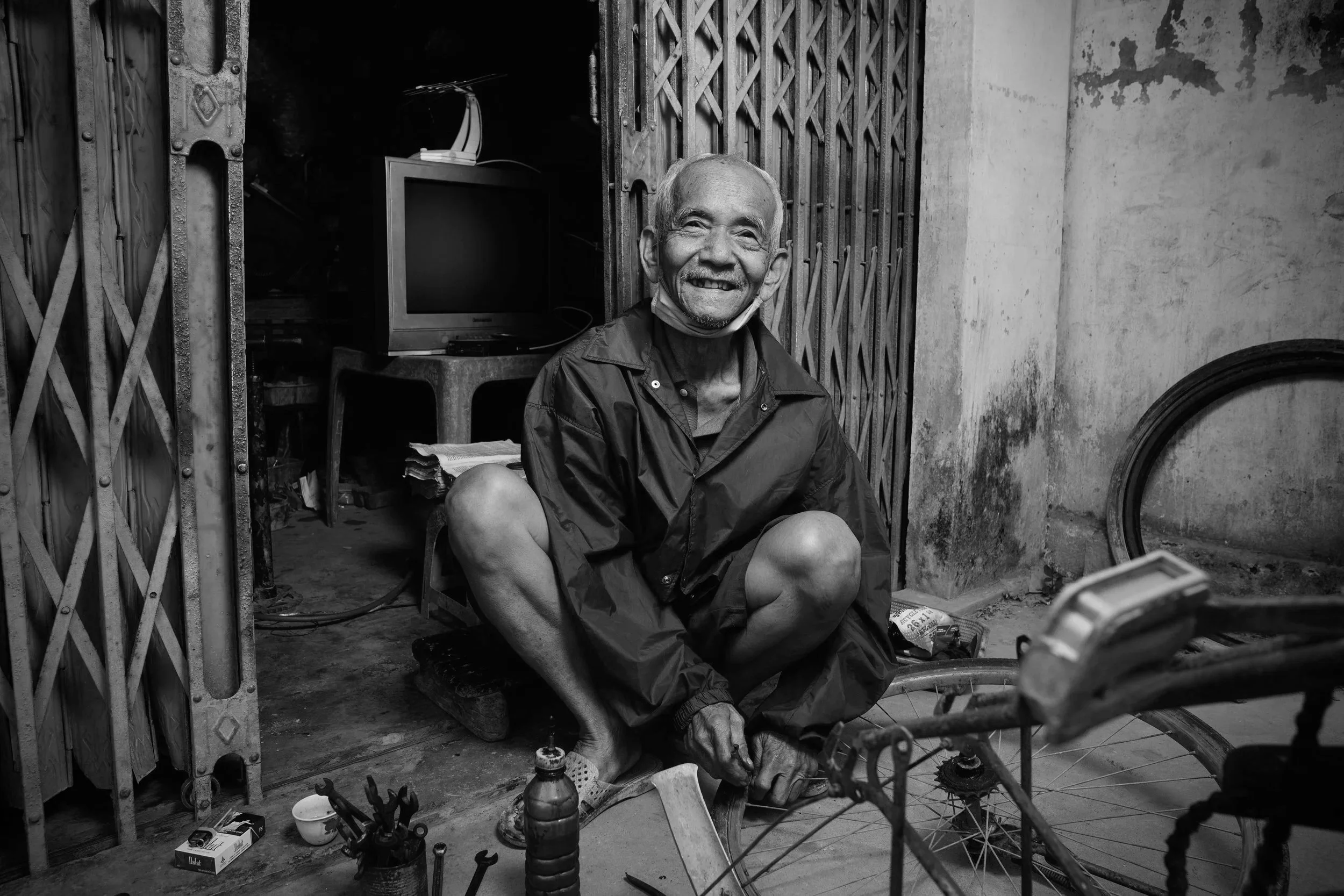An elderly man wearing a blue jacket and shorts, sitting on the ground and working on a bicycle wheel inside a workshop with a blue metal gate, a small TV, and various tools and objects around.