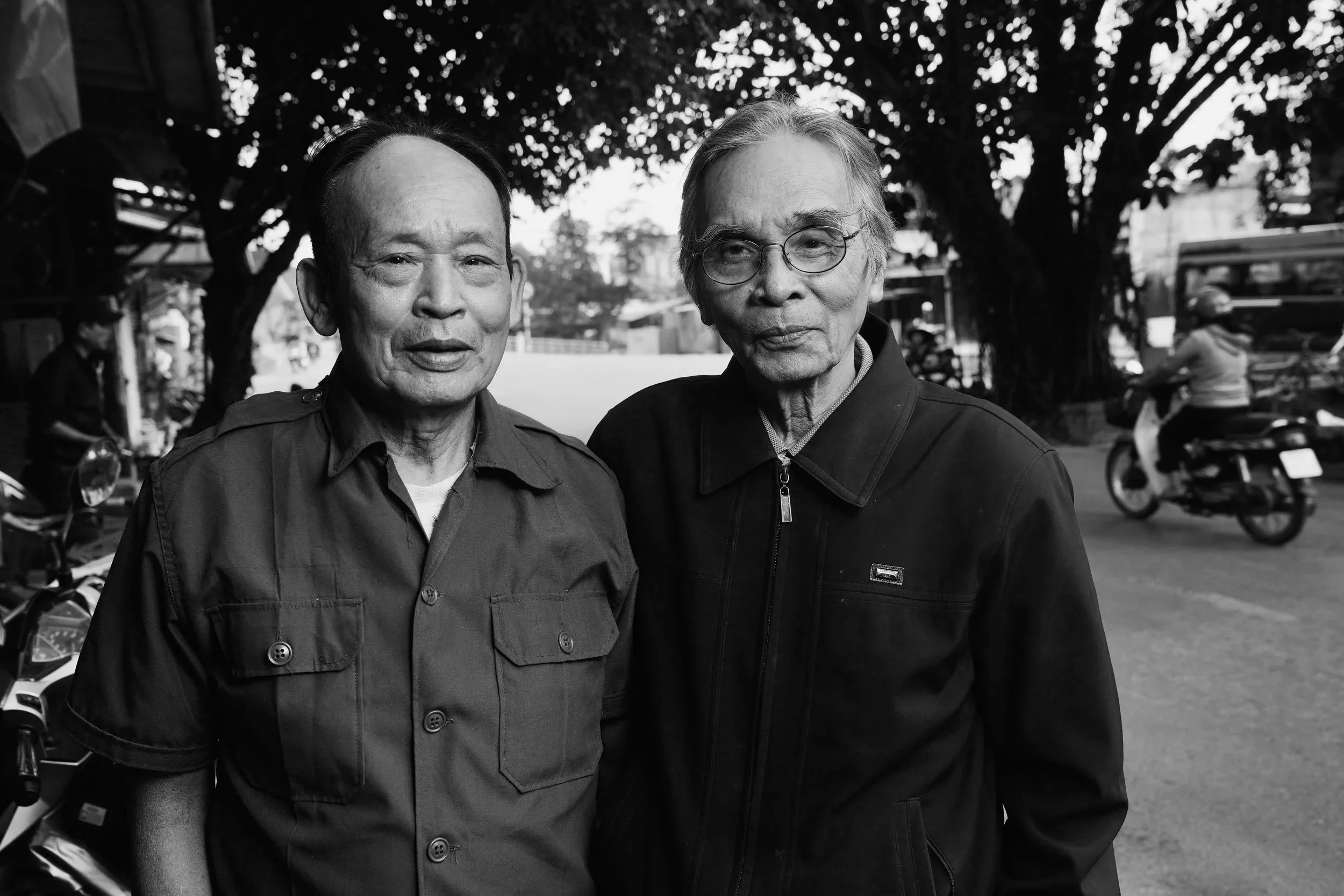 Two elderly men standing outdoors in front of a large tree, posing for the photo, with motorbikes and people in background.
