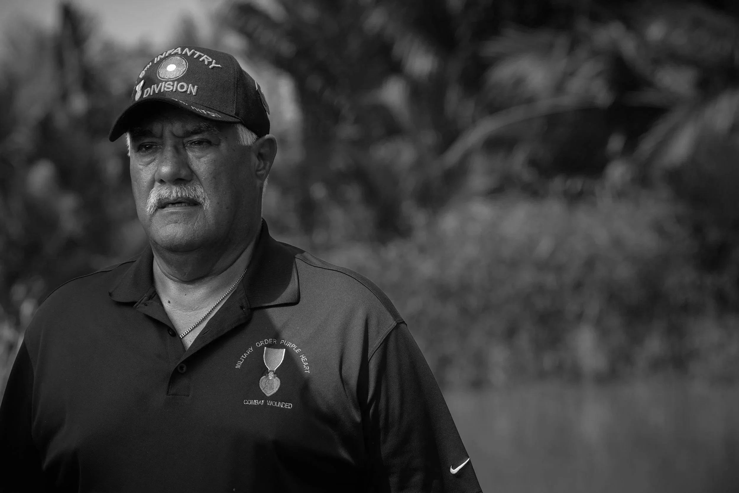A man with a white mustache wearing a black cap with military insignia and the words "Army Infantry Division." He is also wearing a black polo shirt with a purple medal emblem and text that reads "Military Order Purple Heart Combat Wounded." The back