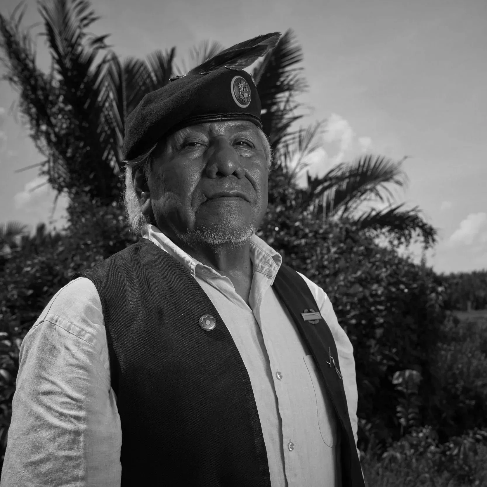 An elderly indigenous man wearing a black feathered headdress, white collared shirt, and black vest stands outdoors amidst greenery, with a blue sky and clouds in the background.