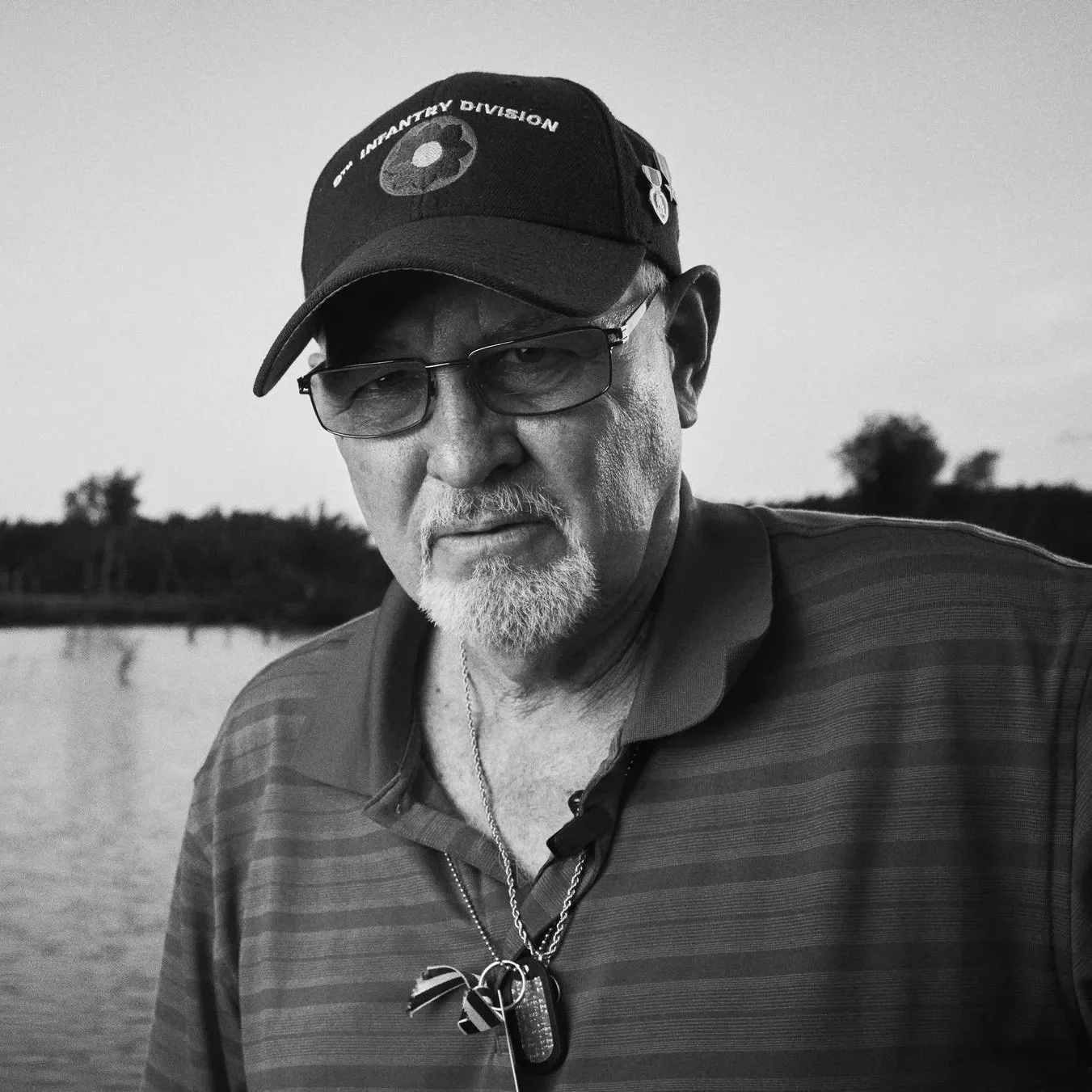 A black-and-white photo of an older man with glasses, a beard, and a mustache. He's wearing a cap with the '77 INFA FATHER DIVISION' emblem, a striped collared shirt with a zipper, and has dog tags around his neck. The background features a body of w