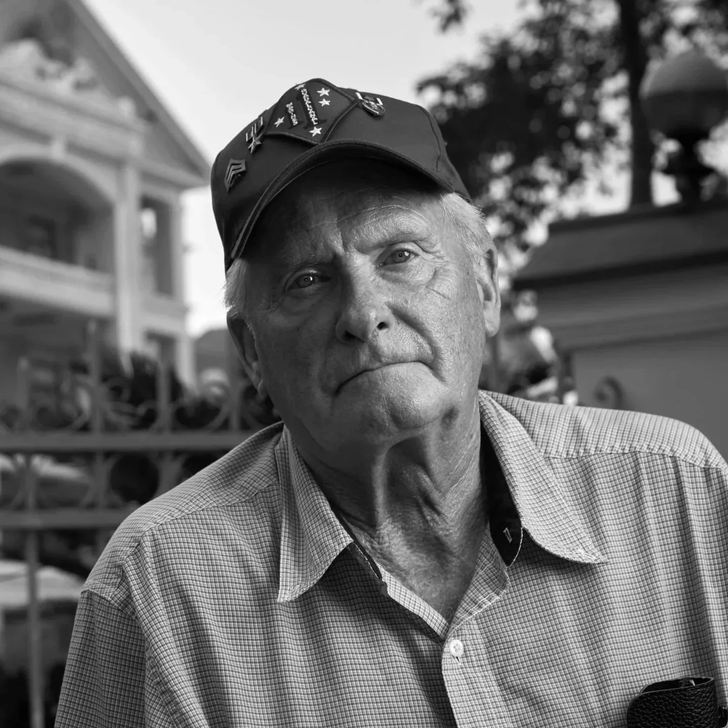 An elderly man with a serious expression wearing a cap with pins and a checkered shirt, standing outdoors with buildings and trees in the background.
