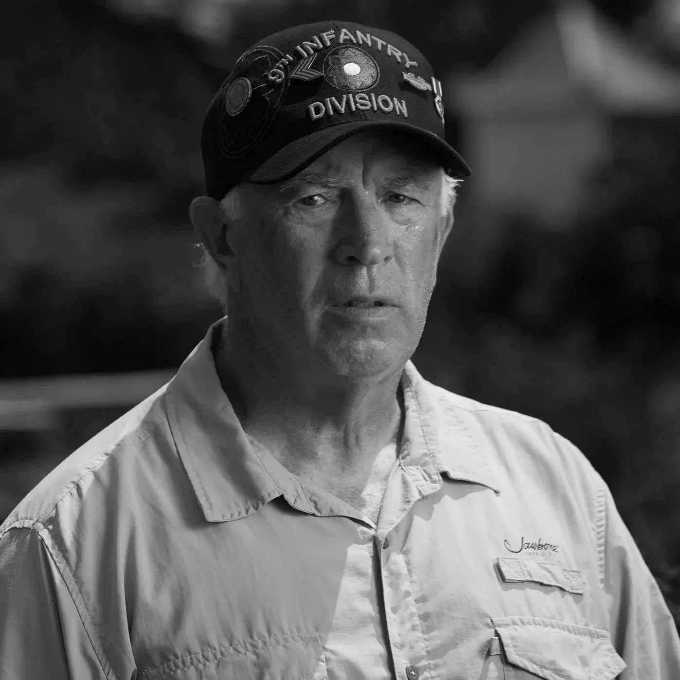 A man wearing a beige button-up shirt and a black cap with patches and the words "Our Infantry Division" visible on it.