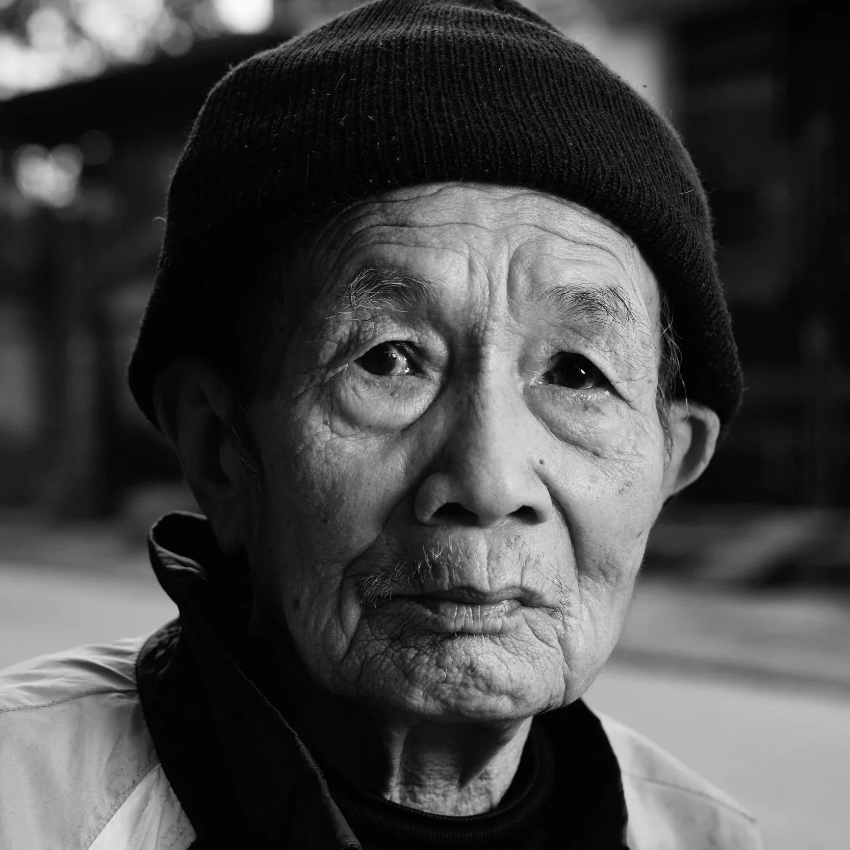 Close-up portrait of an elderly man with a weathered face, wearing a black knit cap, looking directly at the camera, outdoors with blurred background.